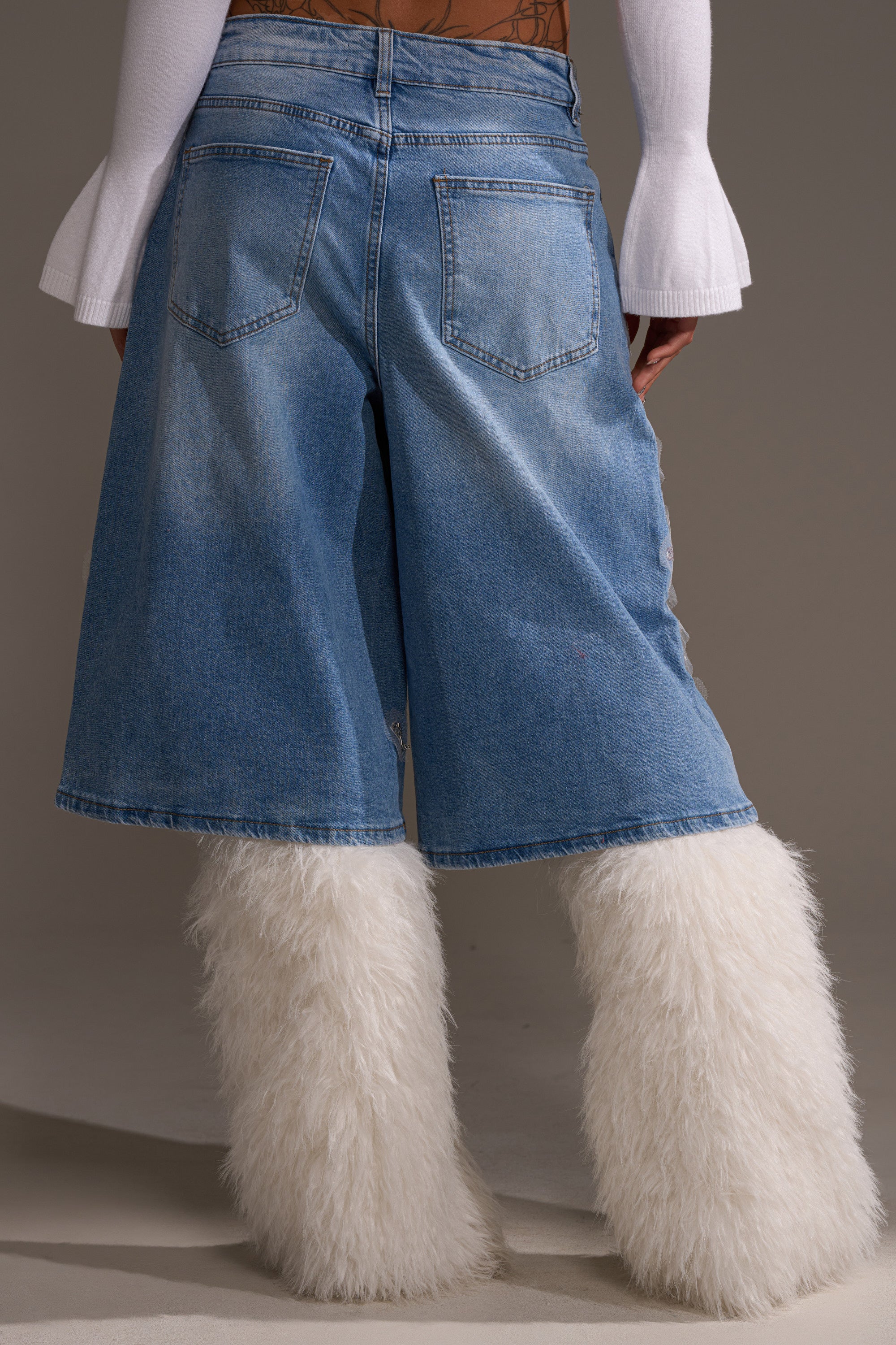 A person shown from the back wears the MADDIE EMBELLISHED BERMUDA SHORT, featuring a light-wash, wide-leg, high-rise fit, paired with fluffy white furry boots and a white long-sleeve top with flared cuffs.