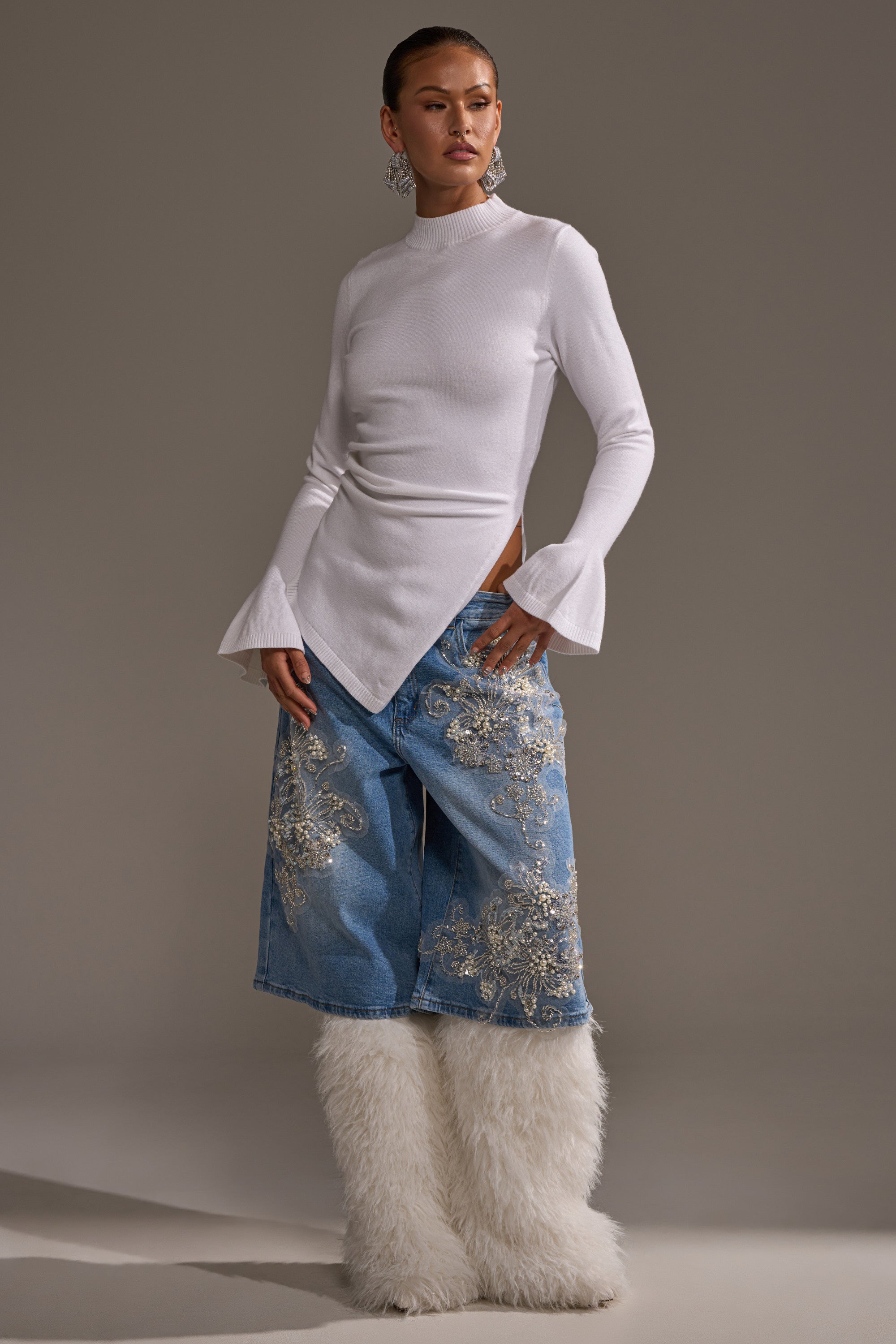 A woman poses in a fitted white top with bell sleeves, the MADDIE EMBELLISHED BERMUDA SHORT in light-wash denim with silver embroidery, tall white fur boots, slicked-back hair, and statement earrings.