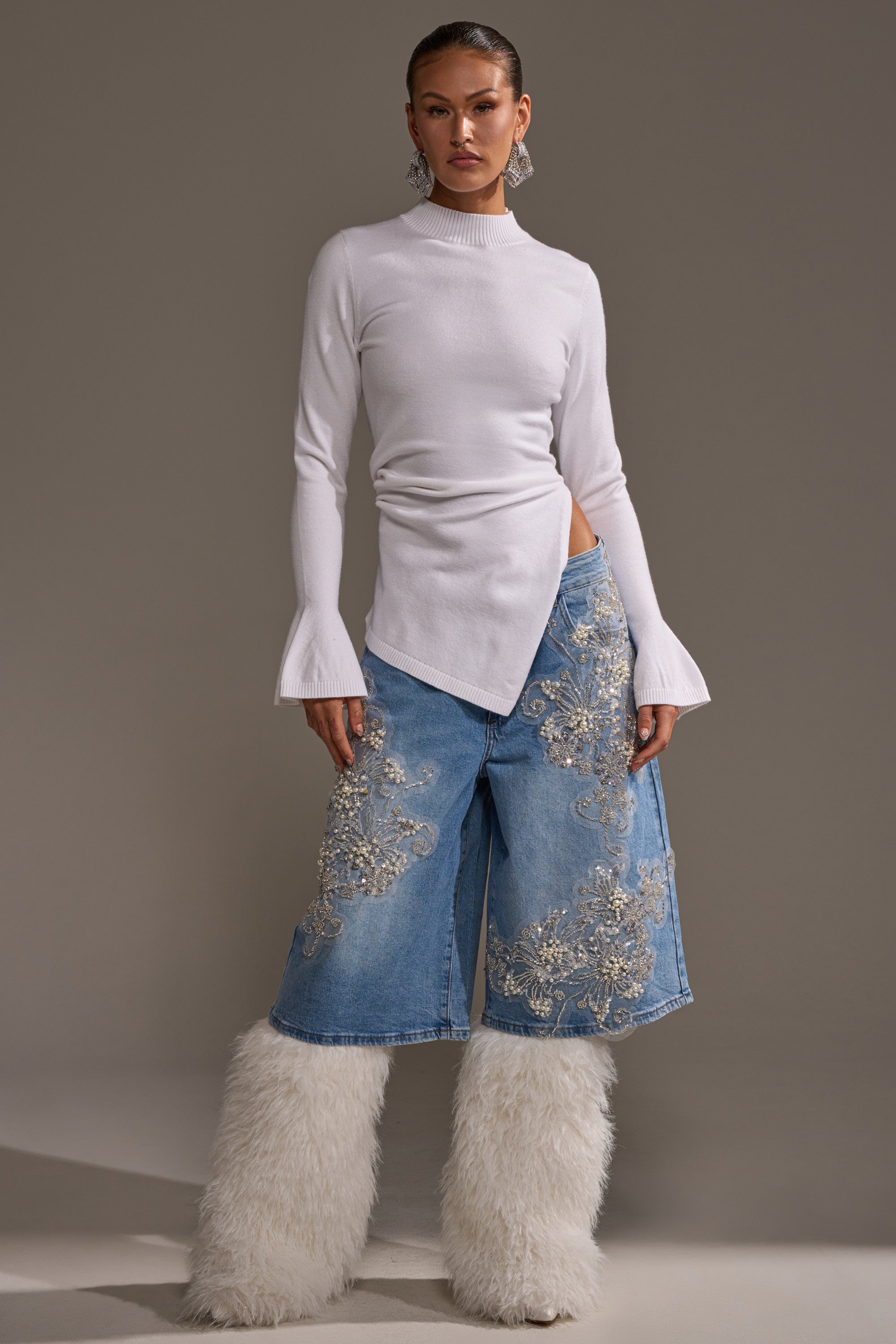 A woman wears the MADDIE EMBELLISHED BERMUDA SHORT—light-wash, high-rise denim with silver embroidery—paired with a white fitted, long-sleeve top, knee-high white furry boots, and large statement earrings.