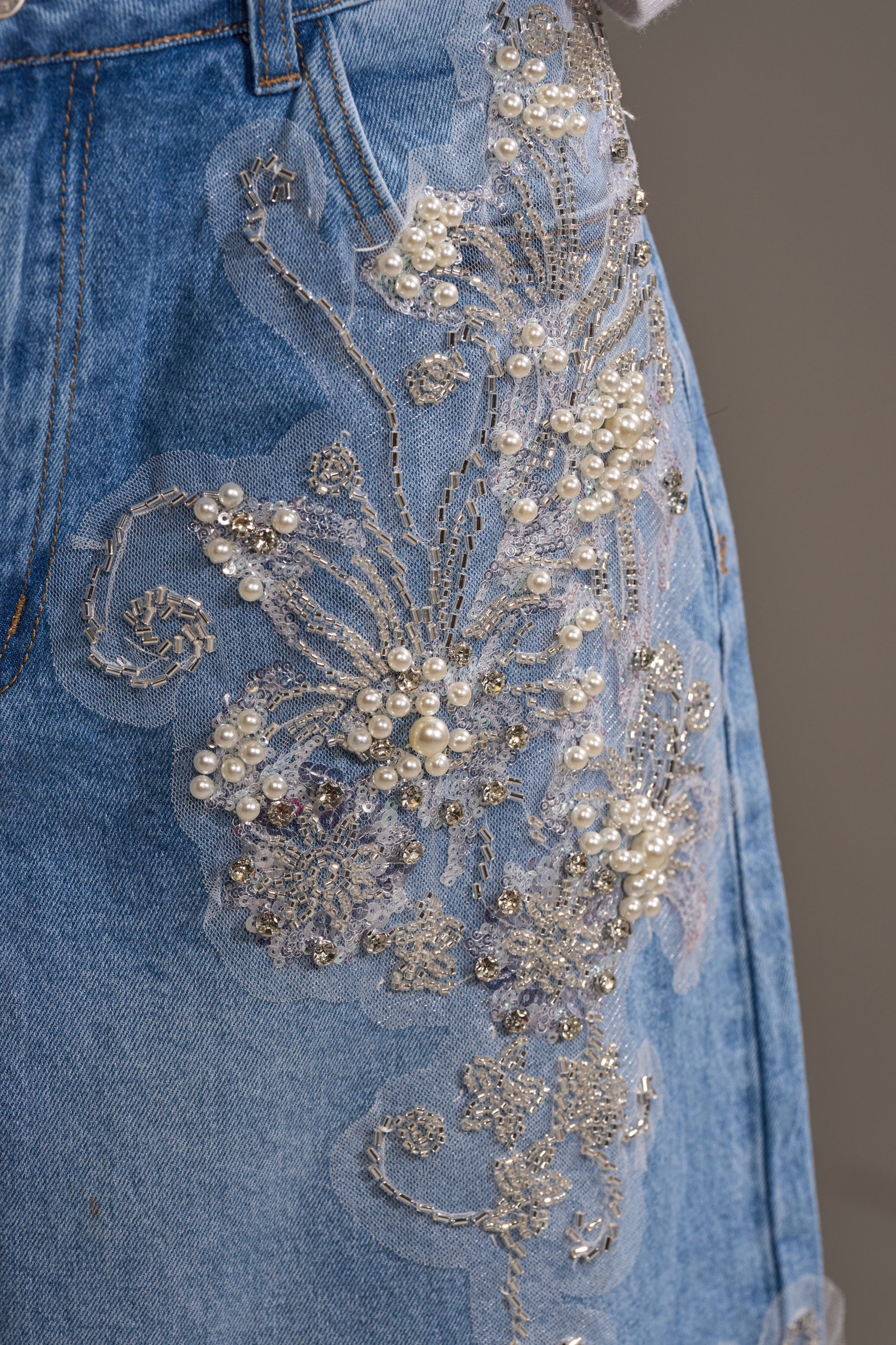 Close-up of the MADDIE EMBELLISHED BERMUDA SHORT in light-wash denim, featuring intricate floral embroidery, silver thread, beads, and pearl accents that form an elegant, textured pattern with a modern high-rise fit.