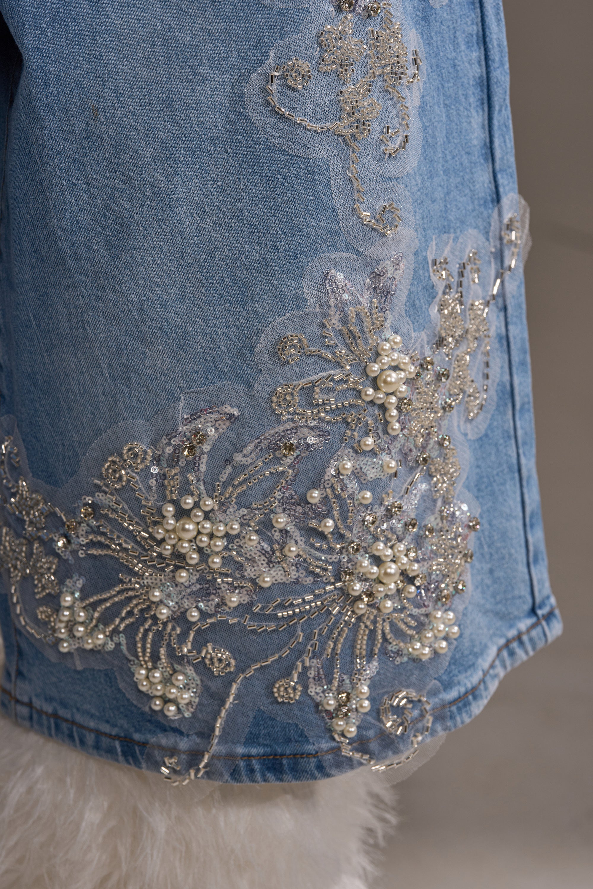 A close-up of the MADDIE EMBELLISHED BERMUDA SHORT in light-wash denim, featuring silver beading, pearls, sequined floral embroidery on sheer fabric, and white fuzzy detail at the high-rise hem.