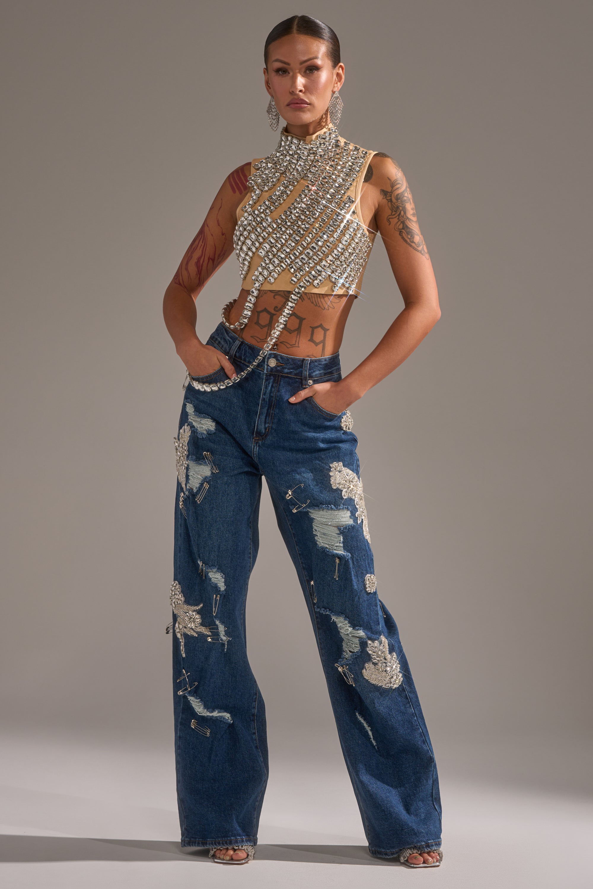 A woman exudes confidence in the LETS GO BABY DISTRESSED DENIM jeans paired with a beaded, jeweled top. With tattoos, large earrings, and slicked-back hair, she showcases a bold and modern style. 1