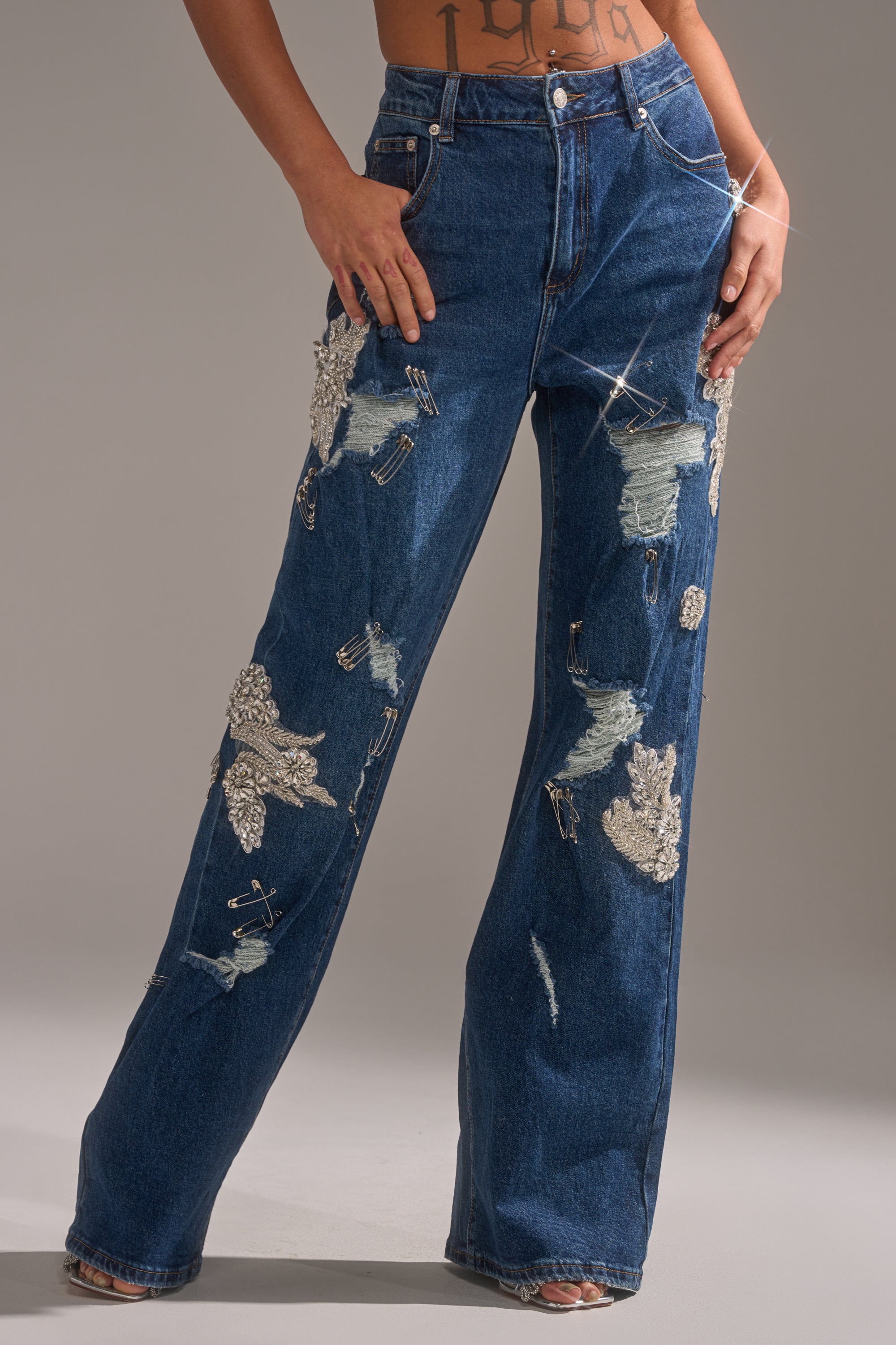 Wearing the LETS GO BABY DISTRESSED DENIM, a person stands with hands on hips in high-waisted, wide-leg jeans featuring sparkling rhinestones, silver leaf patches, and distressed rips. 0