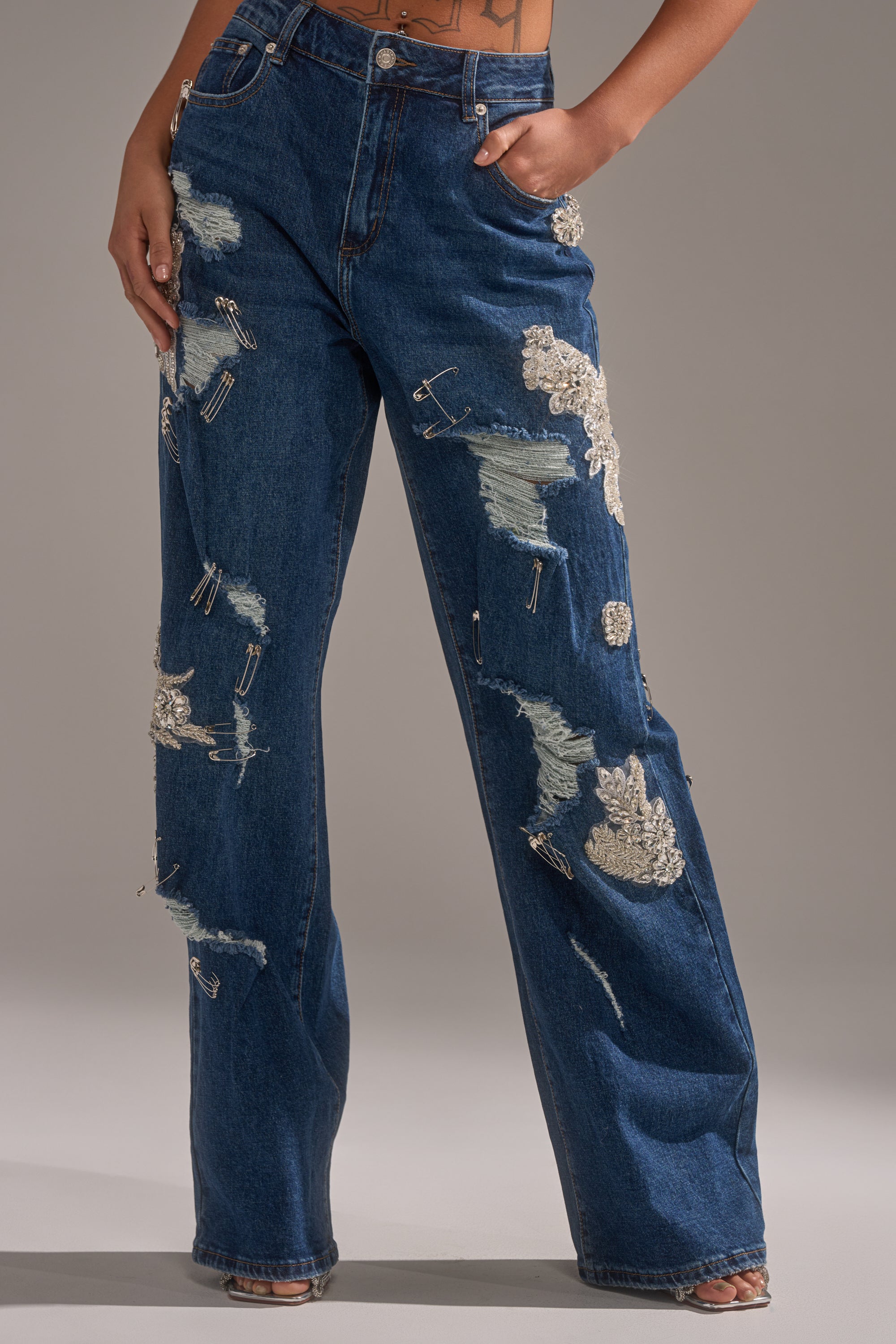 Wearing LETS GO BABY DISTRESSED DENIM—high-waisted, wide-leg jeans with safety pins and silver sequins—a person stands with one hand in the pocket, showcasing this uniquely edgy design. 2