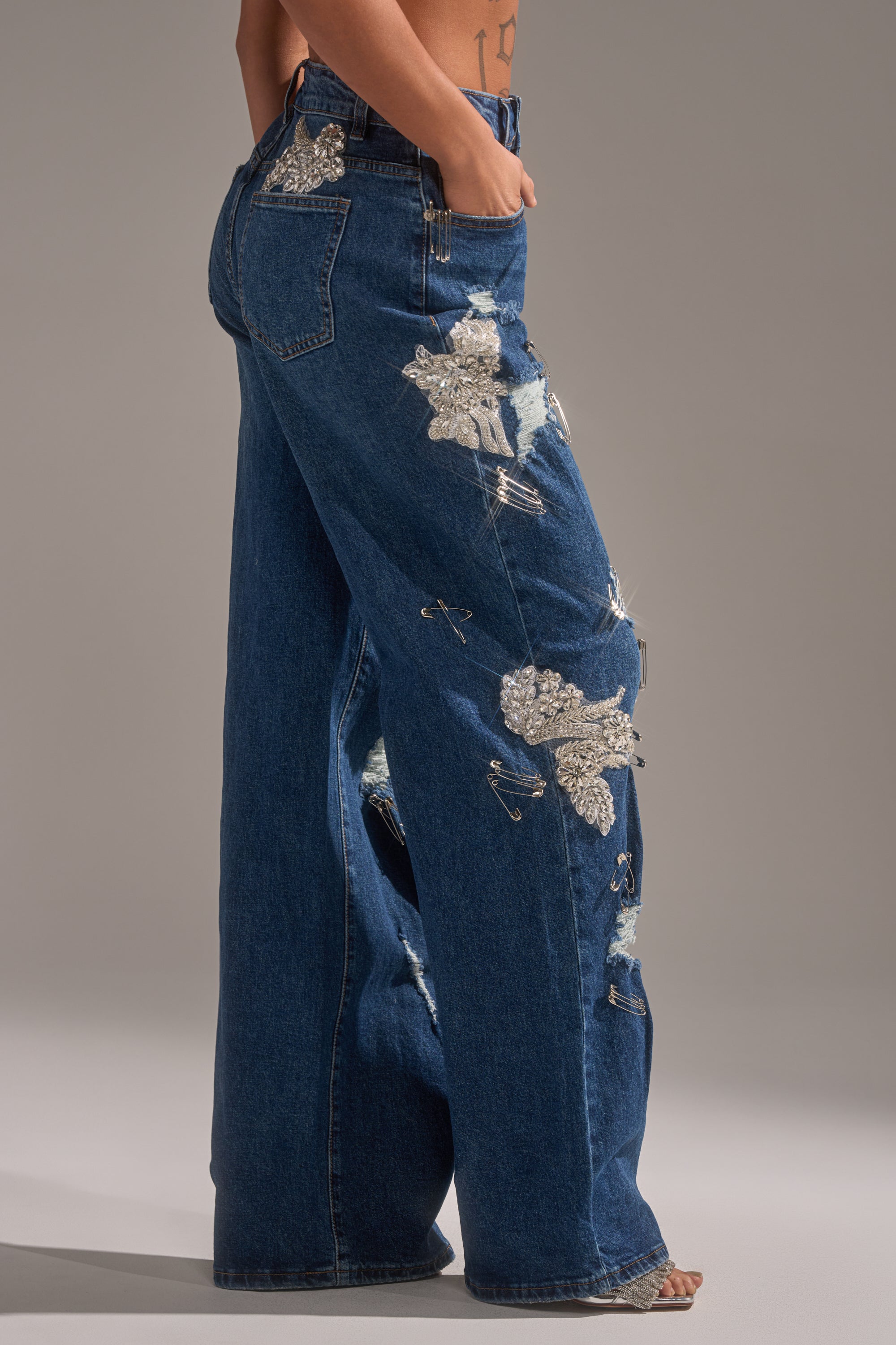 Wearing LETS GO BABY DISTRESSED DENIM—high-rise wide-leg jeans with metallic, beaded details, rips, and safety pins—a person stands sideways with one hand in their pocket, showing a tattoo on their back. 3