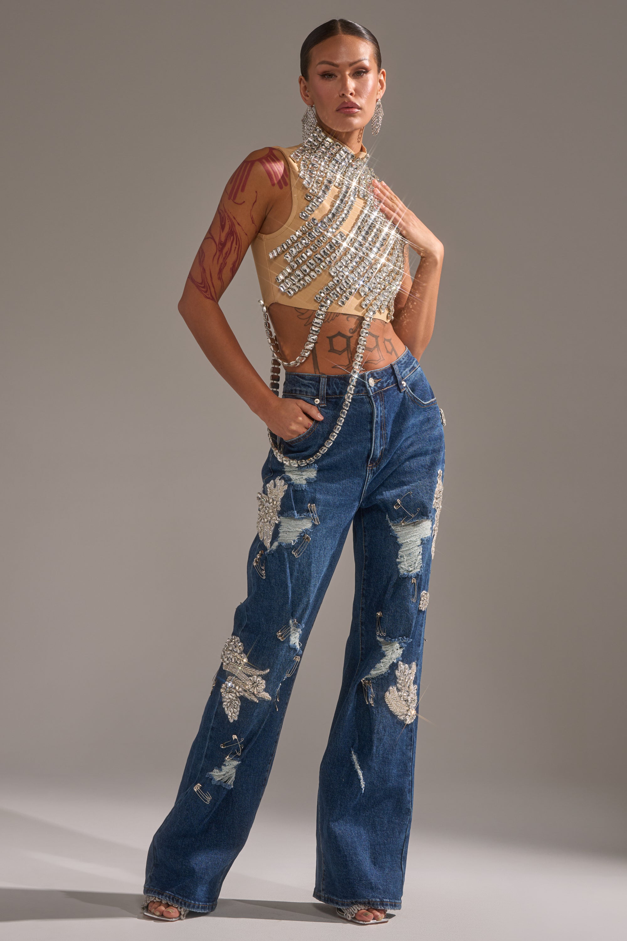 A person with tattoos and slicked-back hair poses confidently against a neutral background, wearing LETS GO BABY DISTRESSED DENIM high-waisted, crystal-embellished jeans and a jeweled sleeveless top with crisscross straps. 5
