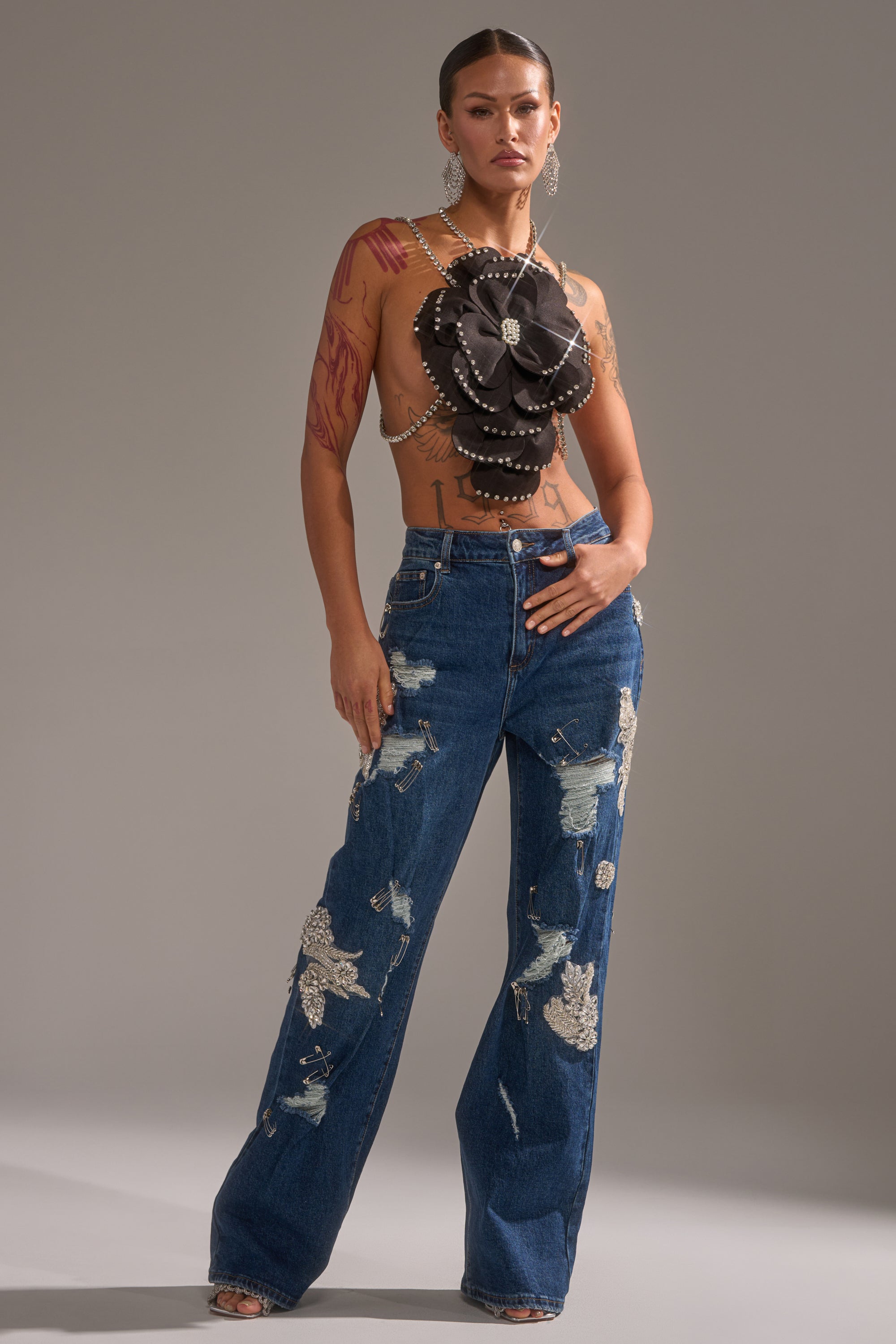 A woman with tattoos, hoop earrings, and a sleek hairstyle poses confidently in LETS GO BABY DISTRESSED DENIM jeans and a dramatic black flower-shaped top adorned with pearls against a plain background. 6