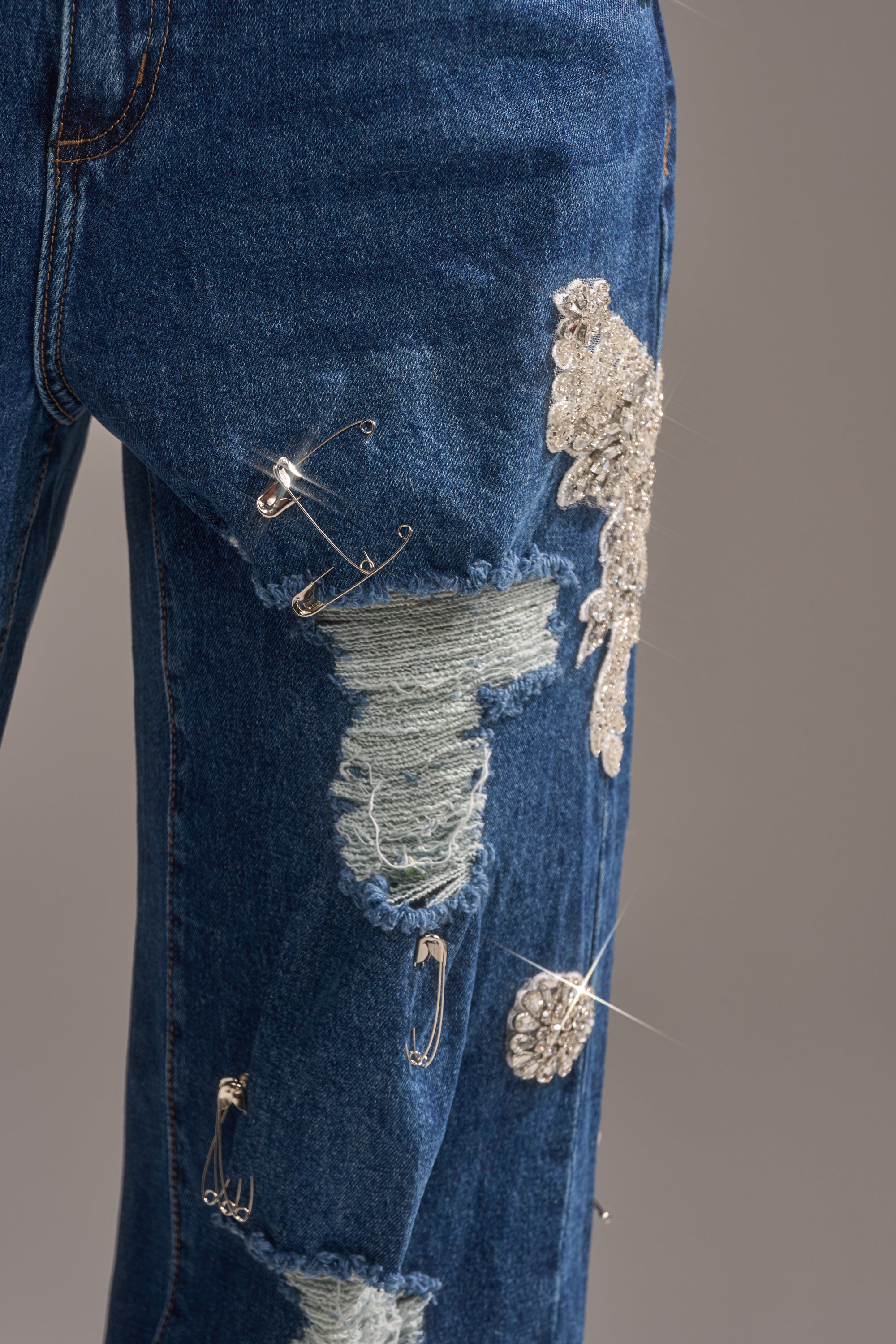 A close-up of LETS GO BABY DISTRESSED DENIM jeans, featuring blue distressed fabric adorned with silver safety pins and sparkling rhinestone embellishments against a plain background. 7