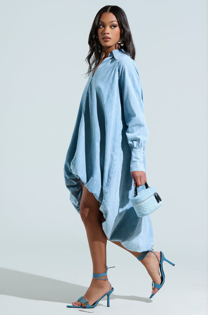 A person wears a loose AMELIA DENIM HIGH-LOW DRESS and matching high-heeled sandals, carrying a small blue handbag. A solid light gray background highlights the casual yet stylish outfit.