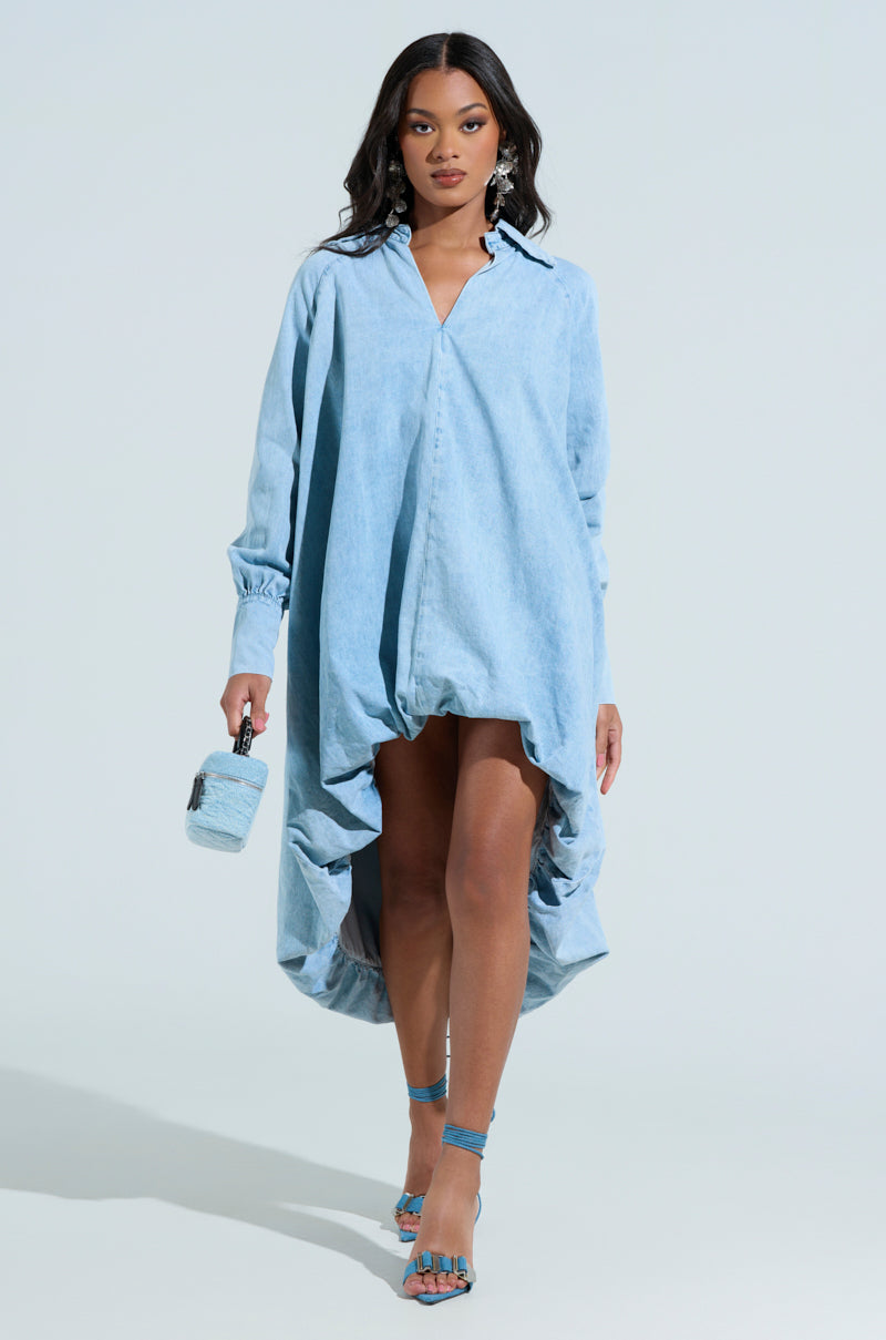 A woman confidently showcases the AMELIA DENIM HIGH-LOW DRESS in light blue, paired with a matching handbag and blue strappy heels. Her hair is styled down, accented by drop earrings against a plain white background.