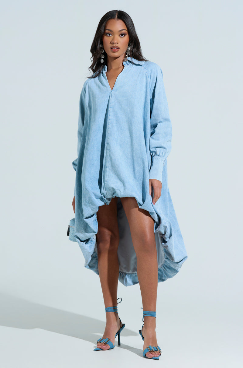 A woman confidently wears the AMELIA DENIM HIGH-LOW DRESS with its oversized fit, paired with matching blue strappy heels. Her long dark hair cascades down her shoulders against a plain light backdrop.