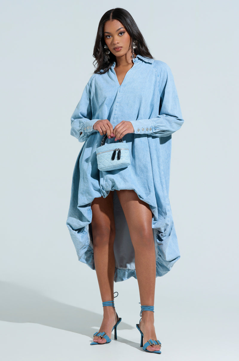 A person with long hair and a confident expression stands against a light background in an Amelia Denim High-Low Dress, pairing it with teal strappy heels and a small textured handbag, highlighting the chic allure of the oversized fit.