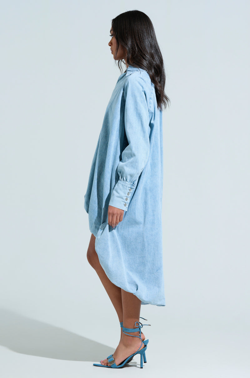 A person with long dark hair stands sideways, highlighting an oversized AMELIA DENIM HIGH-LOW DRESS paired with blue strappy high heels, accentuating the chic high-low hem against a plain light-colored background.