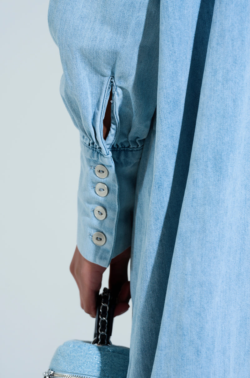 A close-up showcases a person wearing the oversized Amelia Denim High-Low Dress with buttoned cuffs. In their hand is a small denim bag with a chain strap, harmonizing beautifully against a soft gradient background.