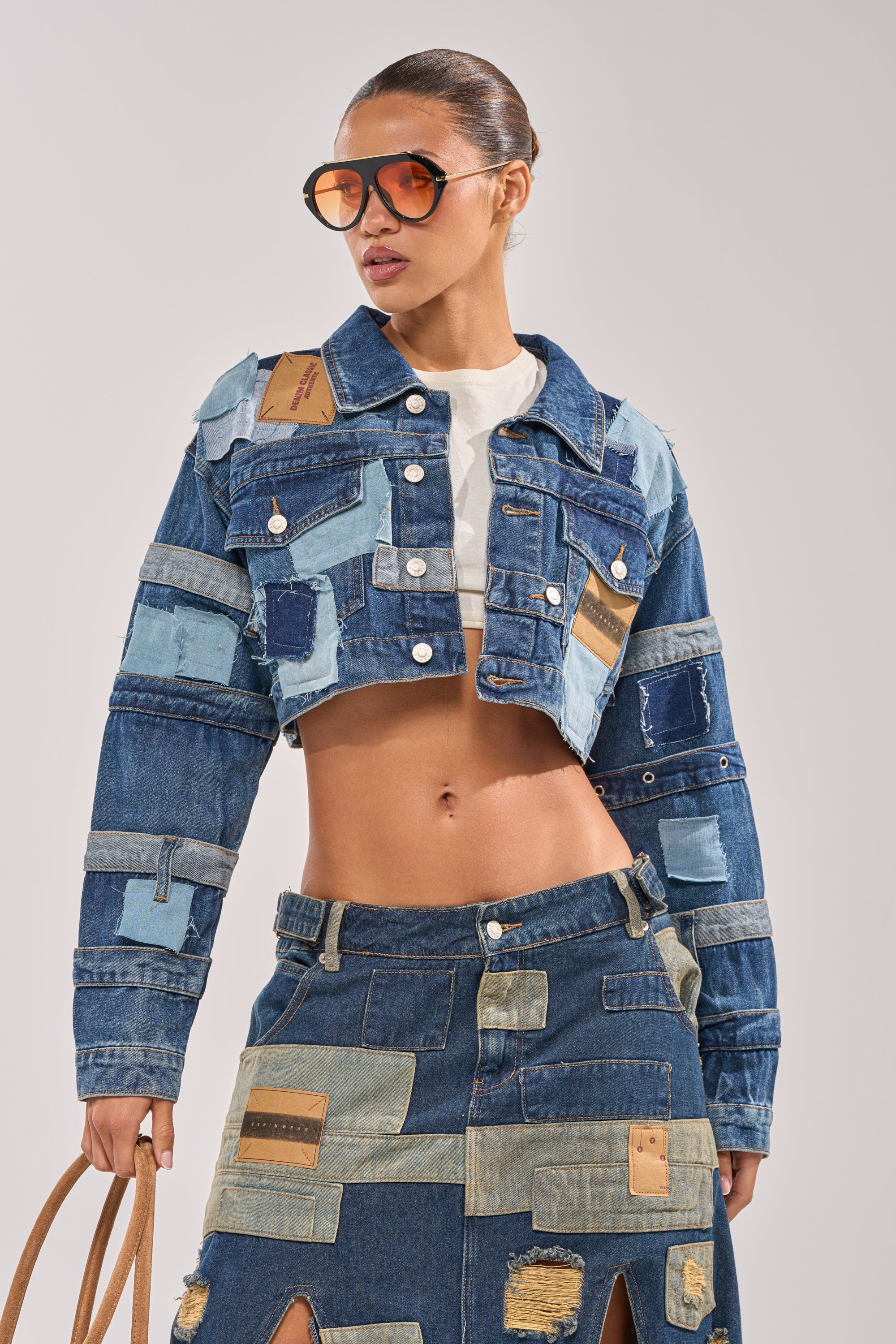 A woman wears oversized sunglasses, the ACT UP PATCHWORK DENIM JACKET, a white crop top, and a matching slit skirt. She holds a tan bag and has slicked-back hair. 0