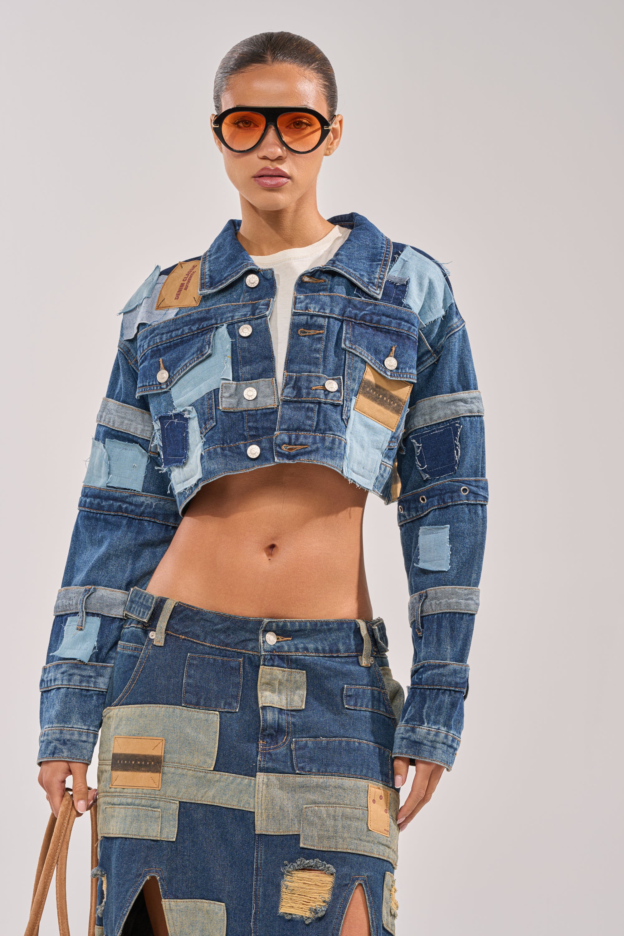 A woman in the ACT UP PATCHWORK DENIM JACKET and matching patchwork slit skirt with large brown sunglasses stands against a plain light background. 1