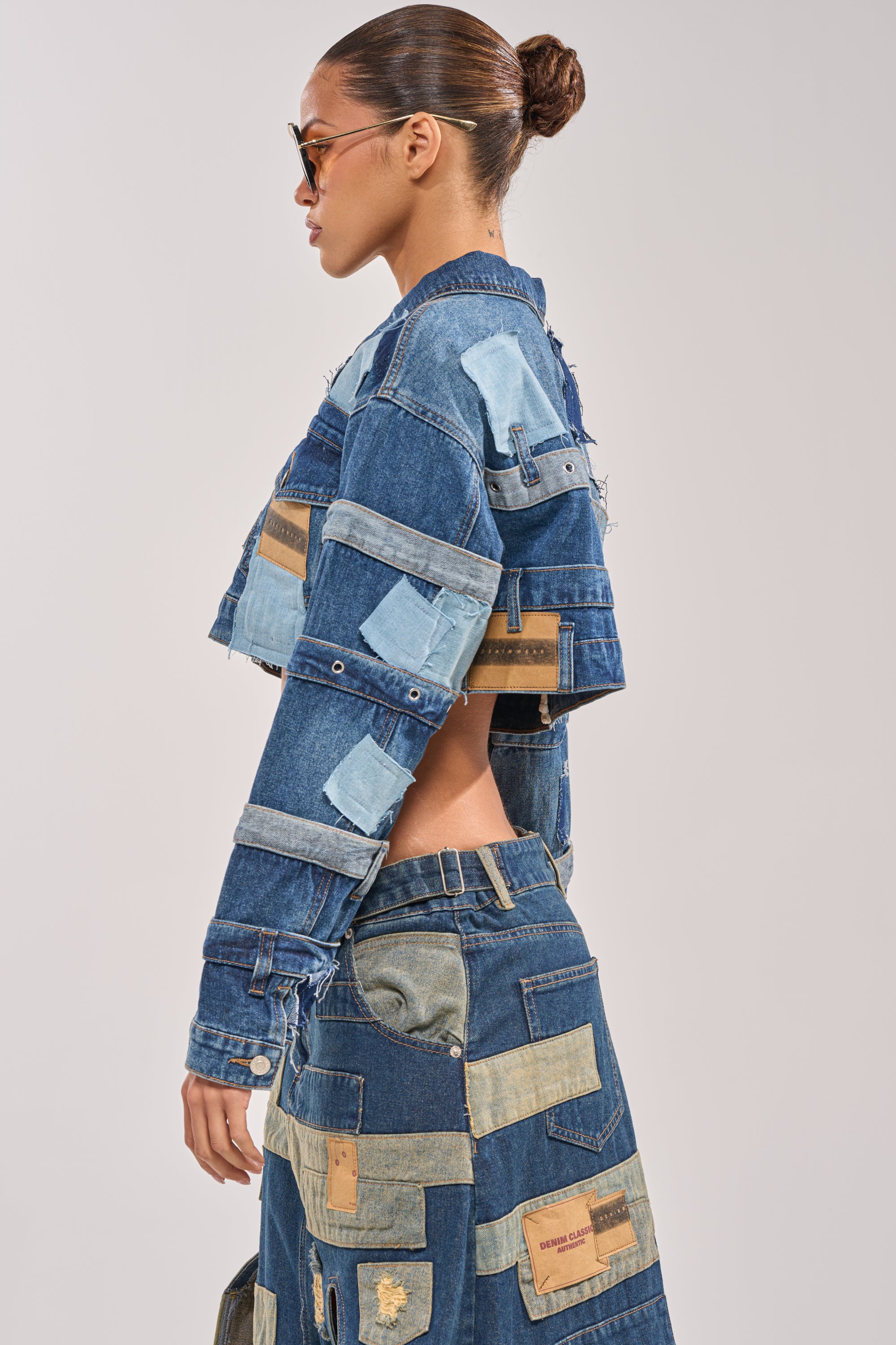 A woman with her hair in a bun wears sunglasses and the ACT UP PATCHWORK DENIM JACKET, paired with a matching patchwork skirt. The outfit features streetwear style with mixed denim shades and textures. She stands in profile against a plain background. 2
