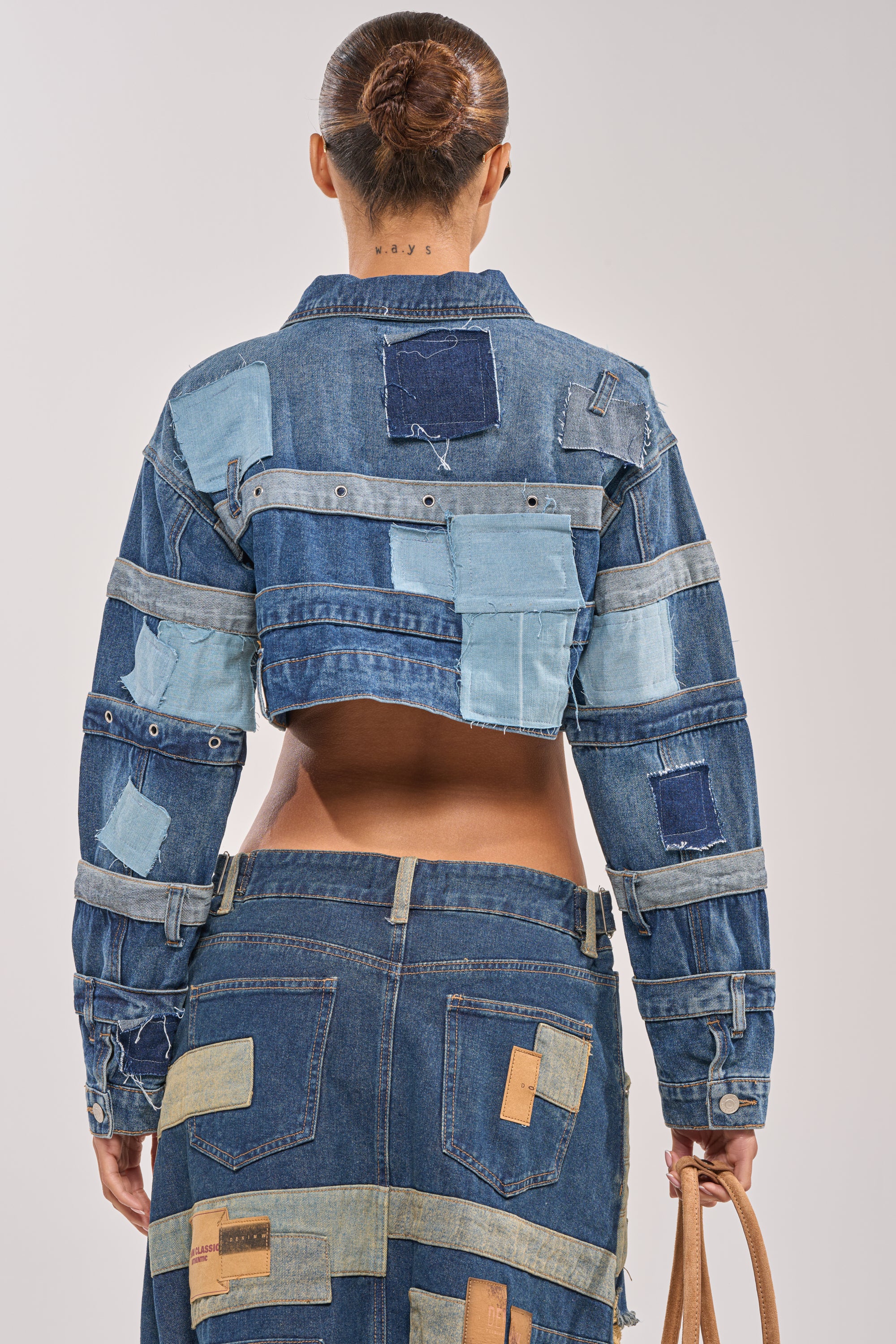 A person stands facing away, wearing the ACT UP PATCHWORK DENIM JACKET and matching skirt in blue tones. Their hair is in a neat bun, and they hold a tan bag, creating a bold streetwear vibe. 3