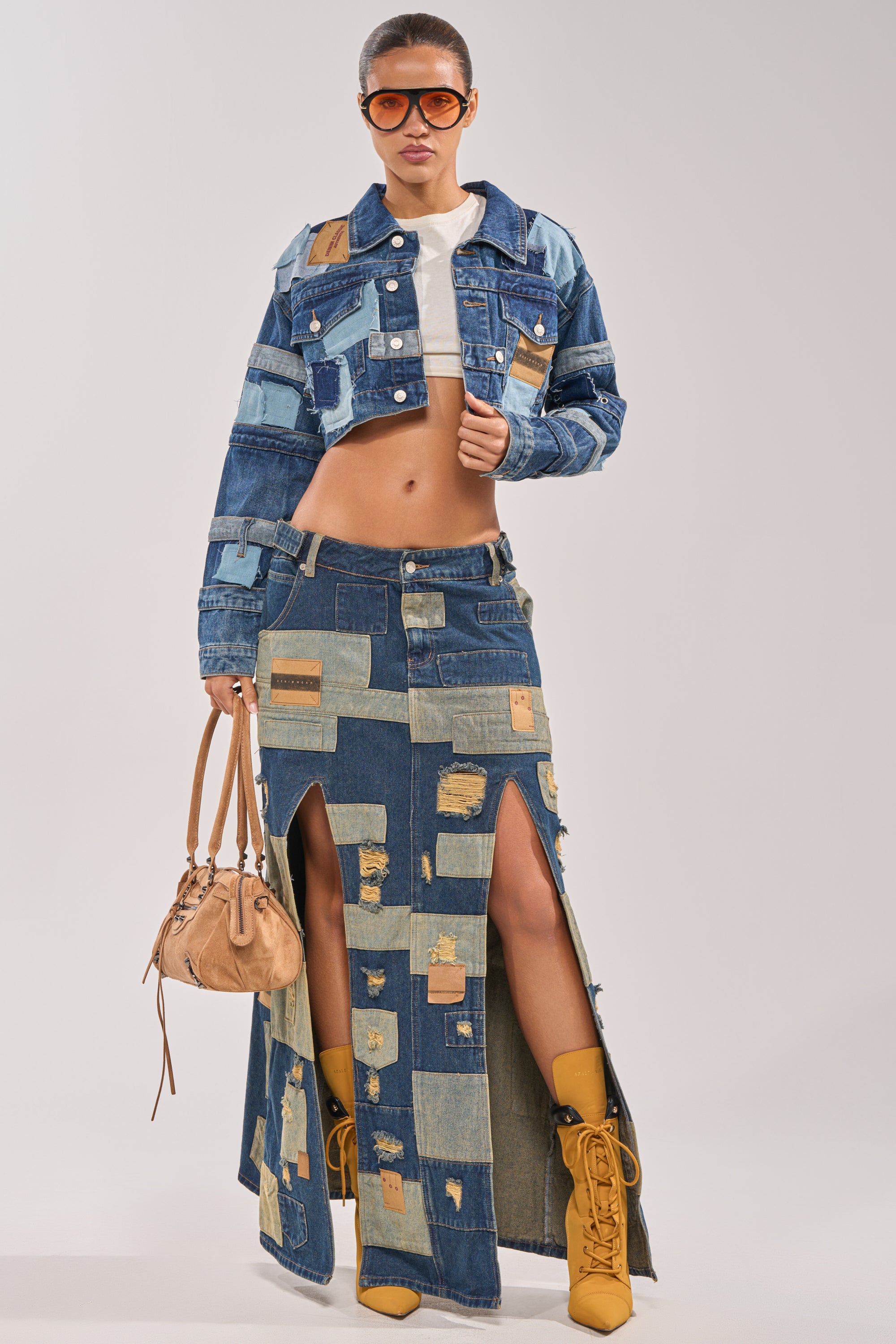 A woman poses confidently against a plain background wearing the ACT UP PATCHWORK DENIM JACKET, matching maxi skirt with front slits, tan boots, oversized sunglasses, and carrying a tan handbag. 5