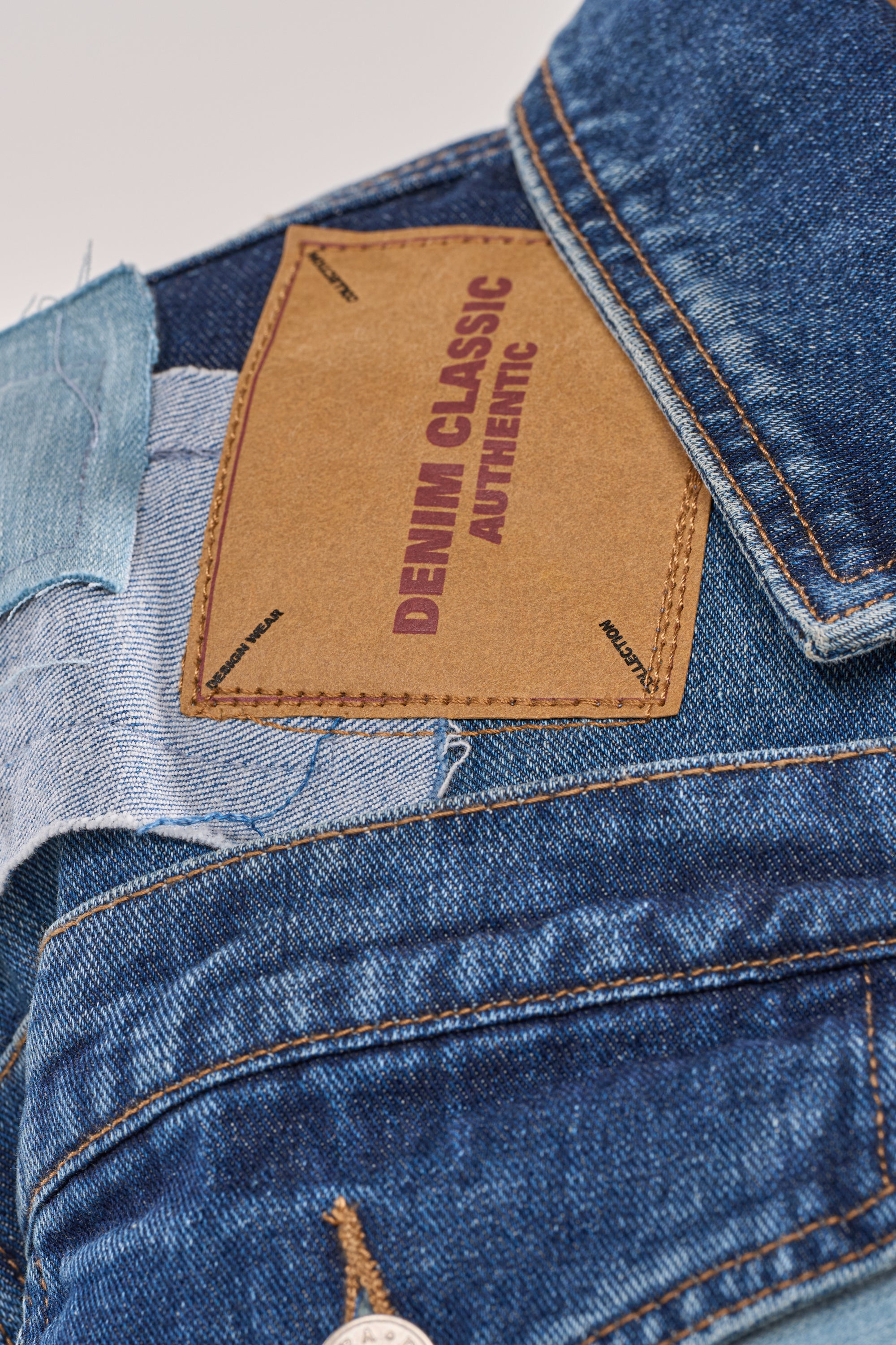 A close-up of blue denim jeans featuring a tan leather patch on the waistband reading "DENIM CLASSIC AUTHENTIC" in bold maroon letters, ideal to pair with your ACT UP PATCHWORK DENIM JACKET. 6