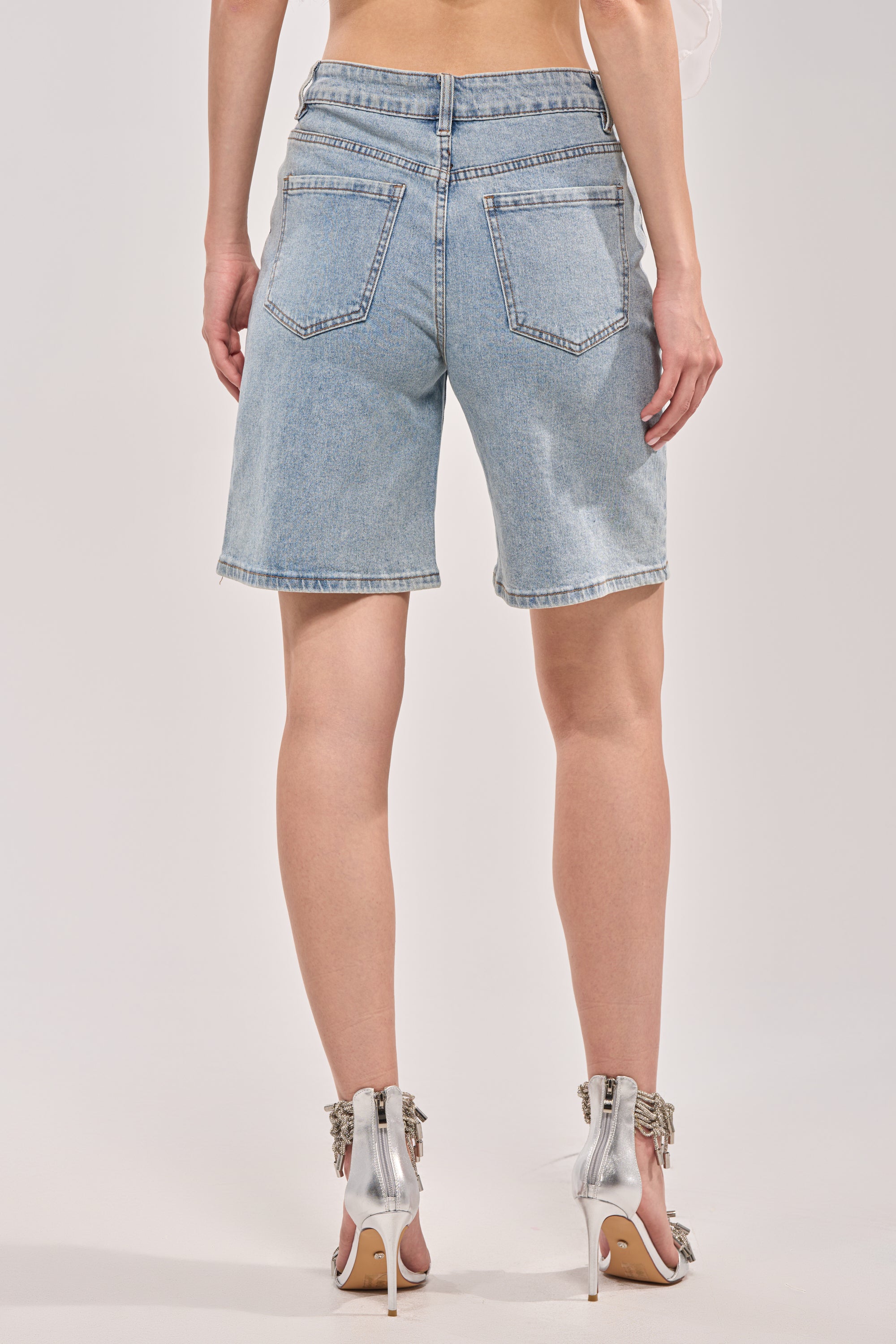 A person stands with their back to the camera against a plain light background, wearing BUTTERFLY QUEEN DENIM SHORTS in light blue with butterfly cutout details and silver high-heeled shoes. 3