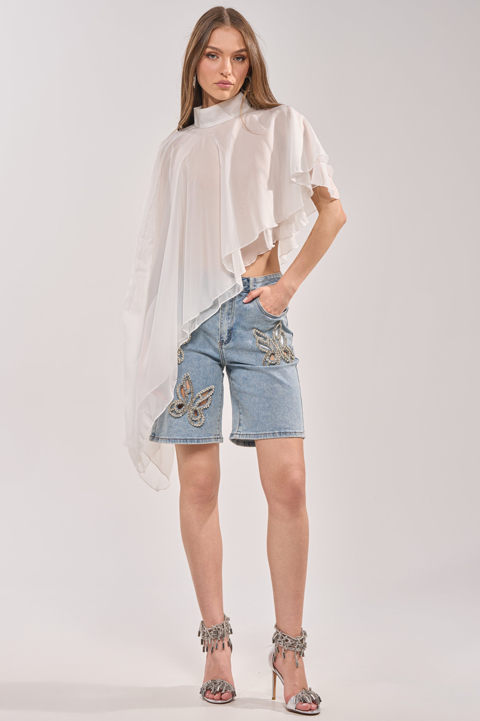 A woman stands against a plain background wearing a sheer, asymmetrical white top, silver high-heeled sandals, and the BUTTERFLY QUEEN DENIM SHORT—knee-length shorts featuring butterfly cutout details. 4