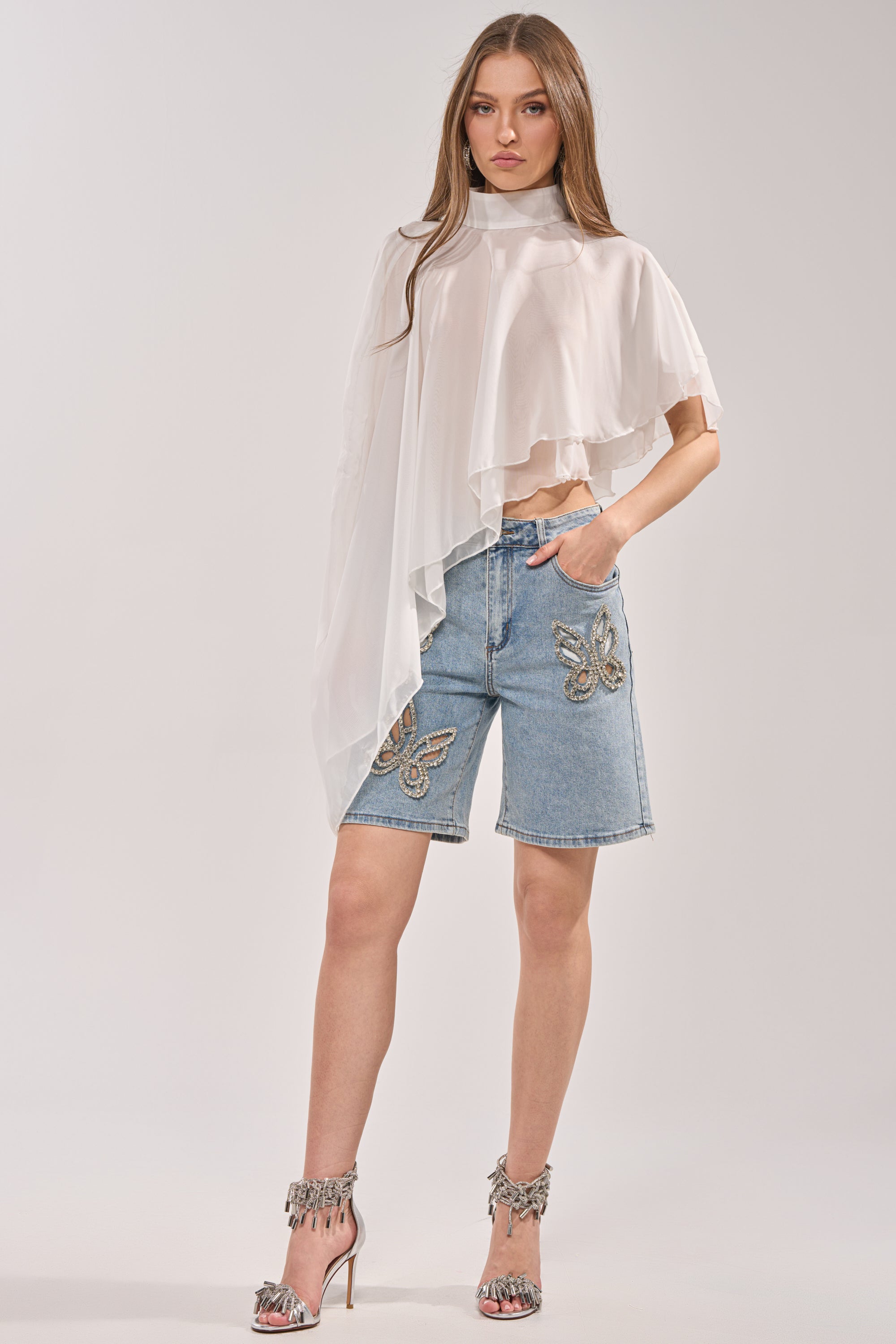 A woman with long brown hair and a neutral expression wears a sheer white asymmetrical blouse, high-heeled fringe sandals, and the BUTTERFLY QUEEN DENIM SHORT featuring butterfly cutouts and rhinestone embellishments. 5
