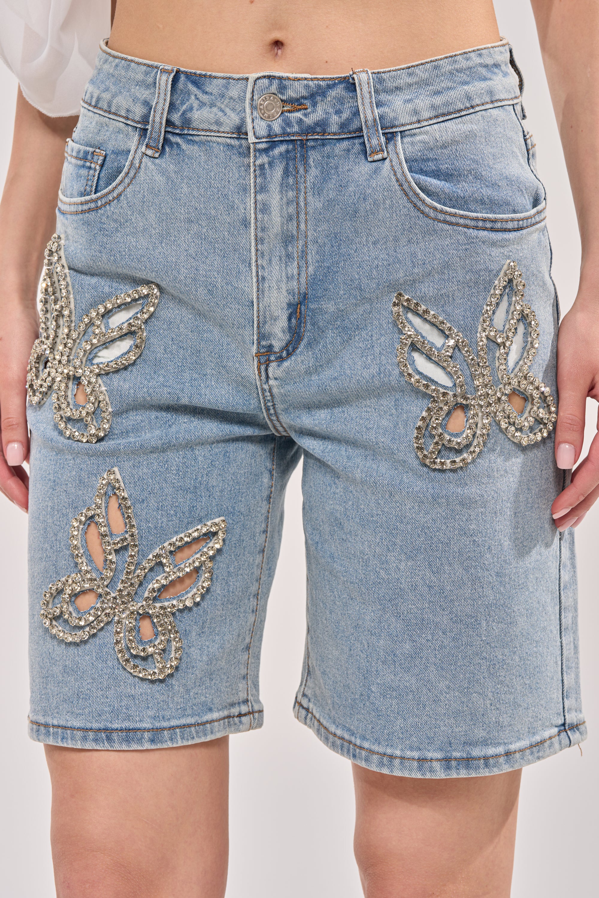 A person wears the BUTTERFLY QUEEN DENIM SHORT—light blue denim shorts with large beaded butterfly appliqués, rhinestone front embellishments, front pockets, belt loops, and a button fly. 6