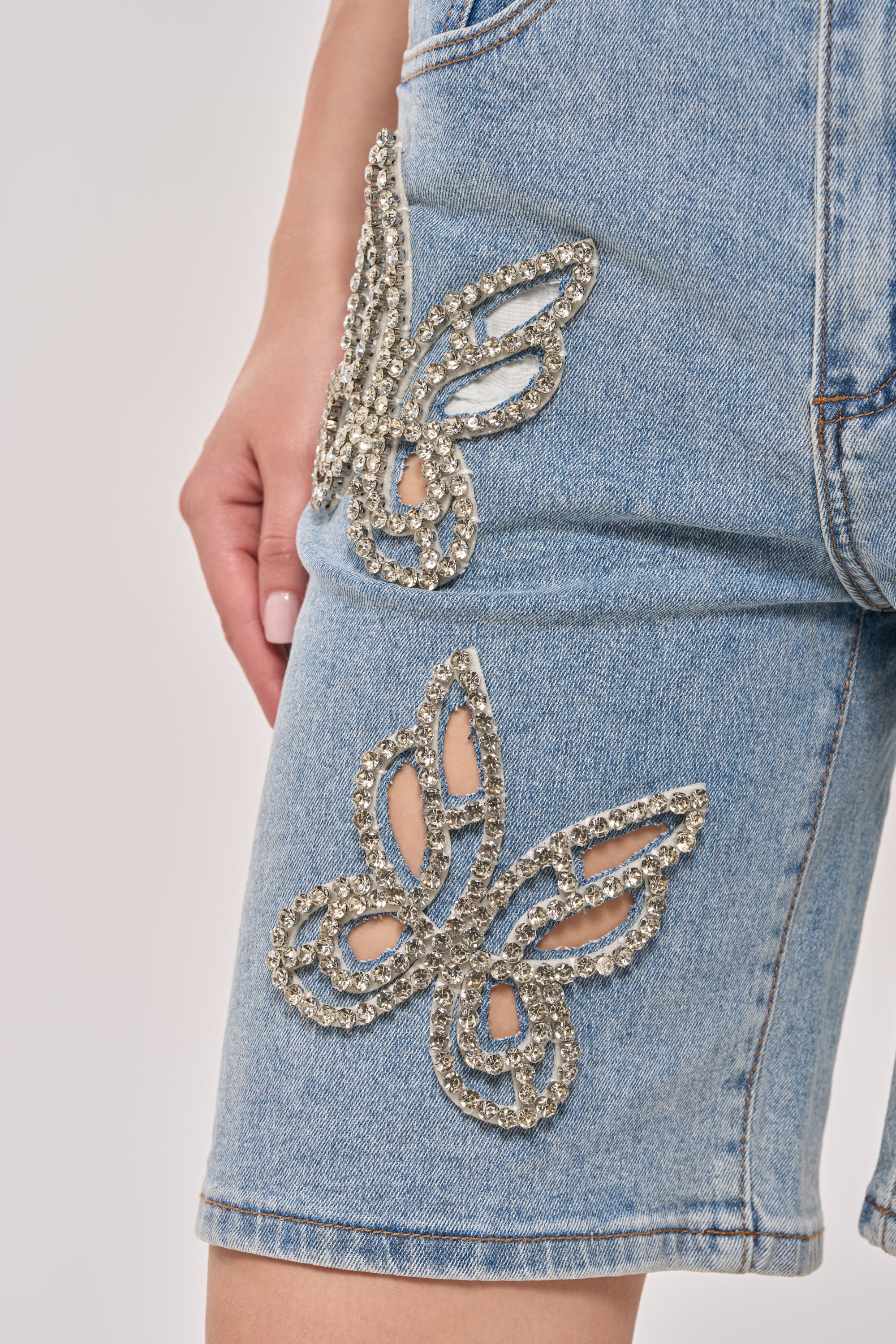 Close-up of a hand resting near the pocket of the BUTTERFLY QUEEN DENIM SHORT, showcasing sparkling rhinestone butterfly embellishments on light blue denim. 7