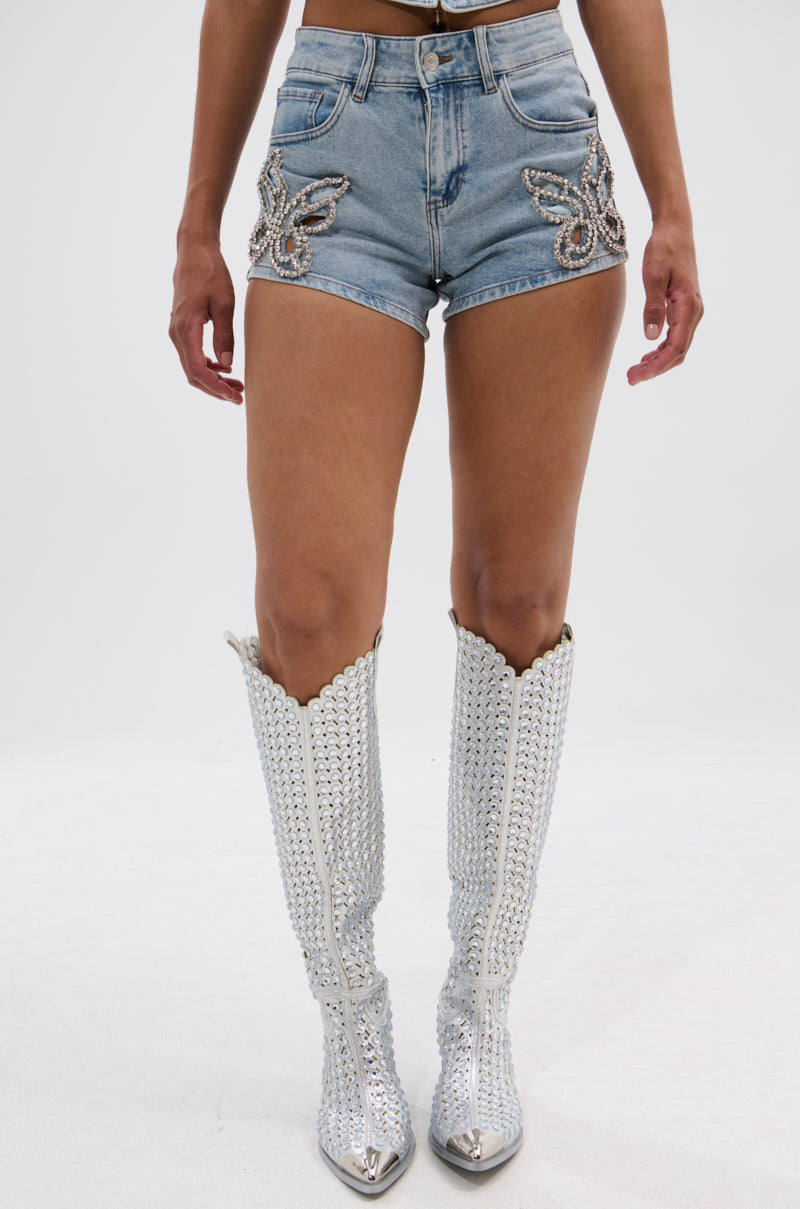 A person models the BUTTERFLY QUEEN DENIM SHORT paired with tall silver studded cowboy boots, standing against a plain white background. 1