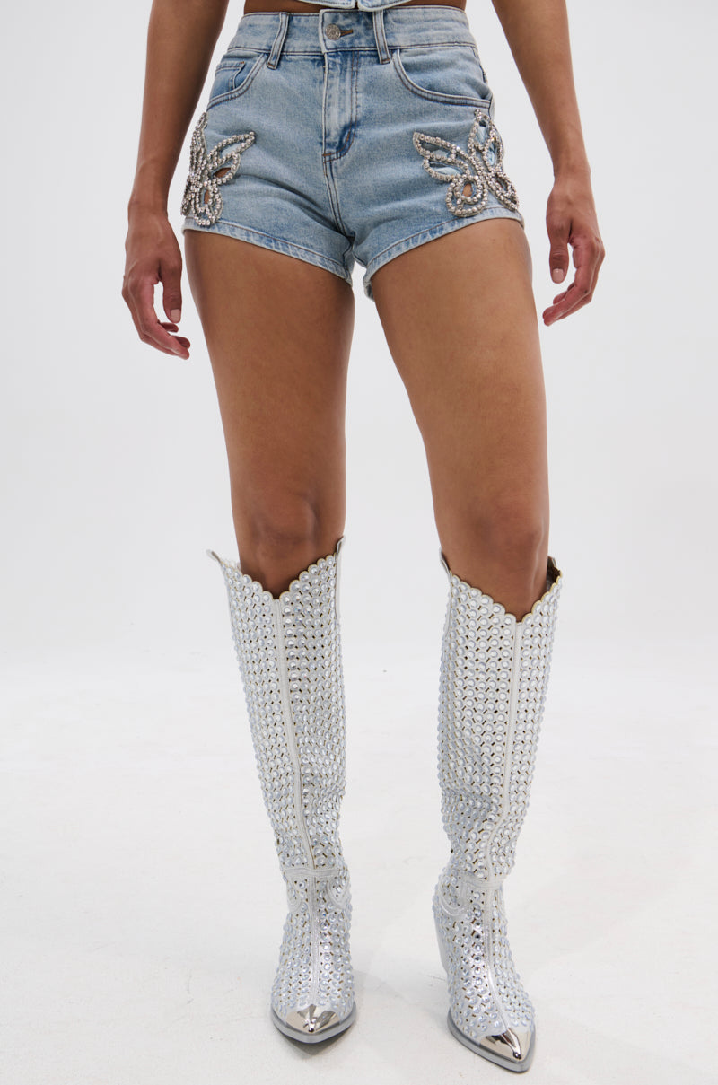 A person wears the BUTTERFLY QUEEN DENIM SHORT—high-rise, butterfly sequin denim shorts—paired with tall silver-studded cowboy boots, standing against a plain white background. 2