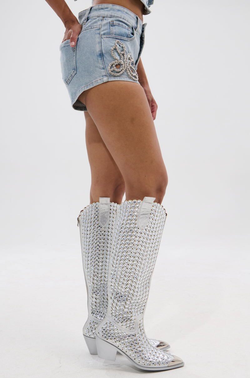 A person wearing the BUTTERFLY QUEEN DENIM SHORT and tall, shiny silver cowboy boots stands against a plain white background. 3