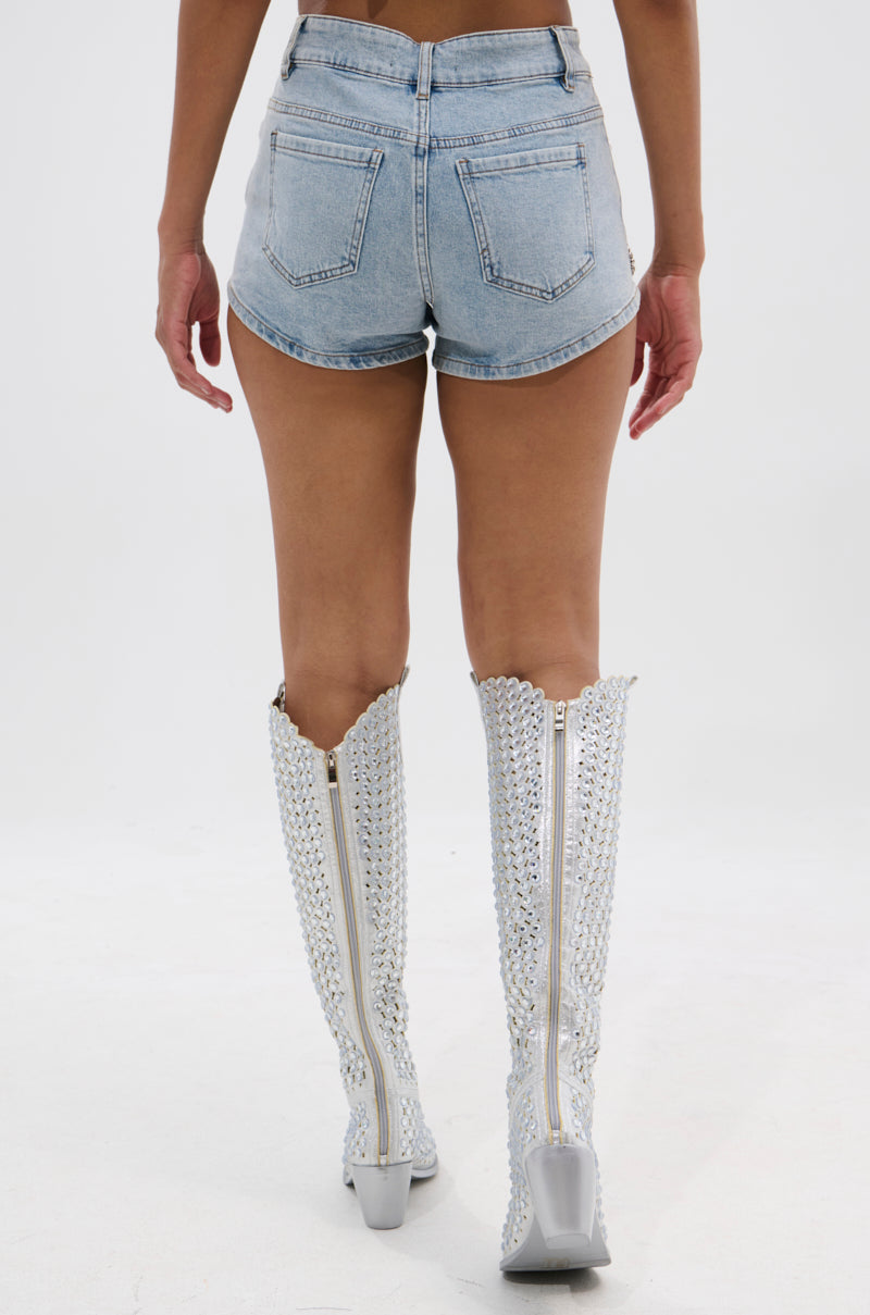 Someone stands facing away against a plain white background, wearing the BUTTERFLY QUEEN DENIM SHORT in light blue and knee-high, silver, textured boots with zippers. 4
