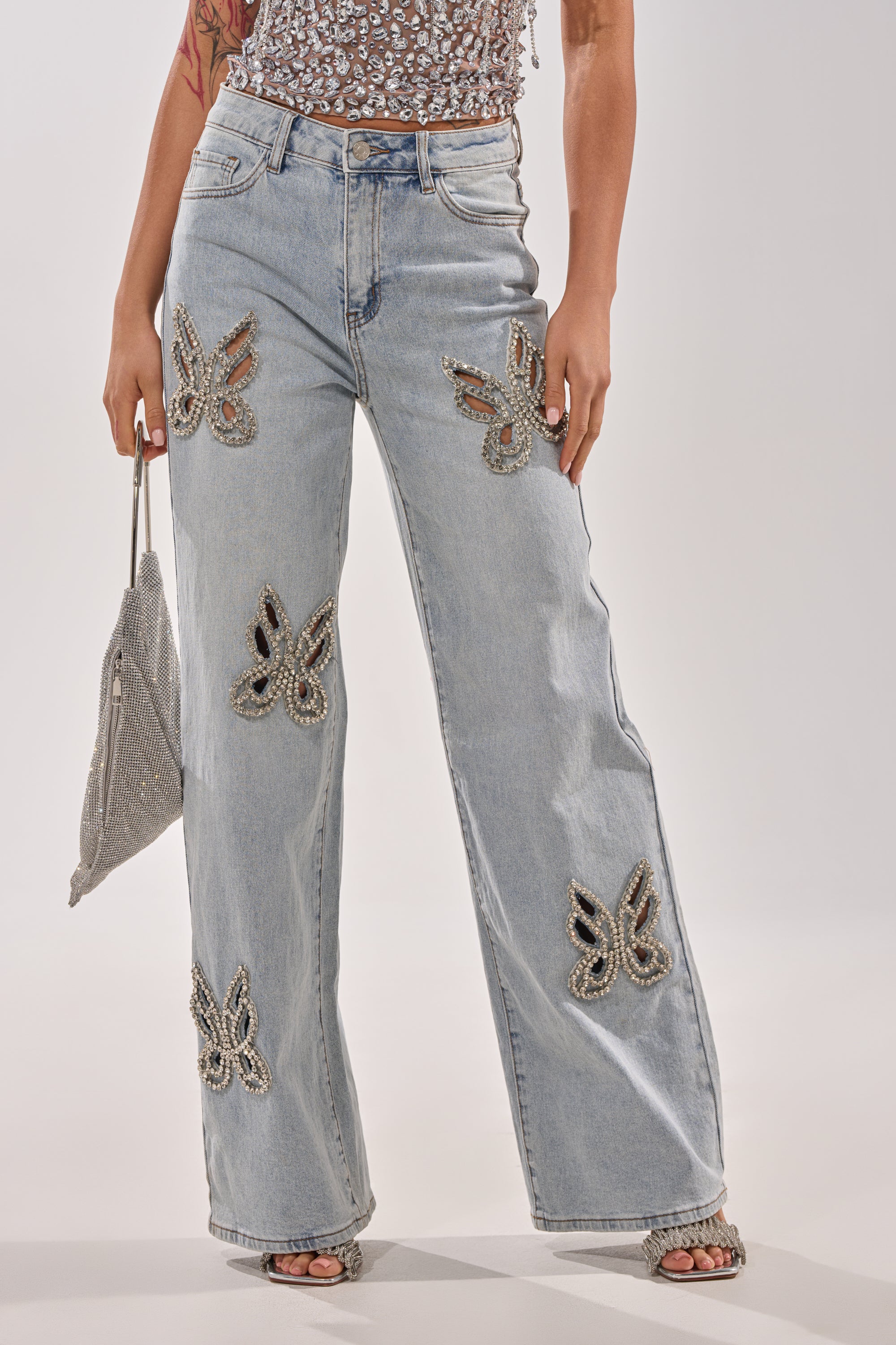 Wearing RHINESTONE BUTTERFLY CUTOUT DENIM JEANS with beaded butterfly details, a sheer embroidered top, and holding a sparkly silver bag. Only the outfit's lower body is visible. 0