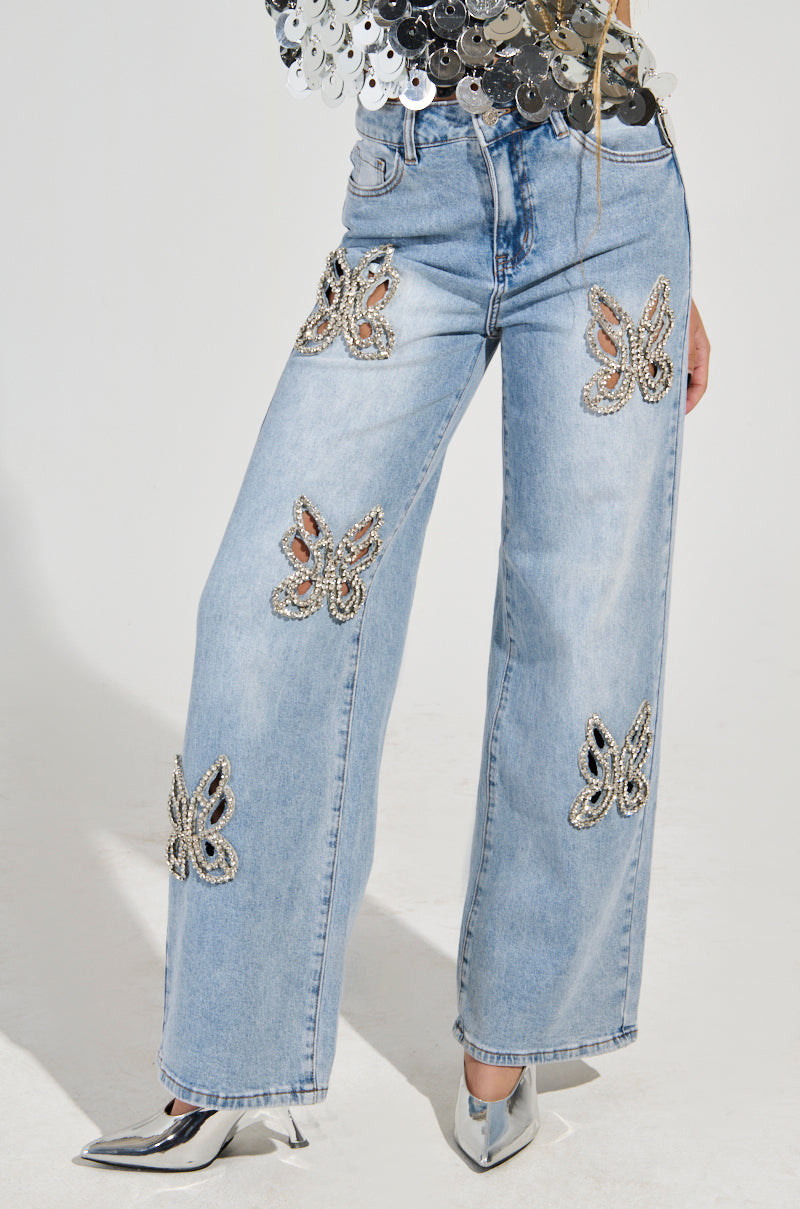 A person stands in Y2K-inspired denim, showcasing the RHINESTONE BUTTERFLY CUTOUT DENIM JEANS in light blue wide-leg style. Their silver pointed shoes glimmer beneath a top with a shiny, reflective texture. The background is a plain, neutral color.