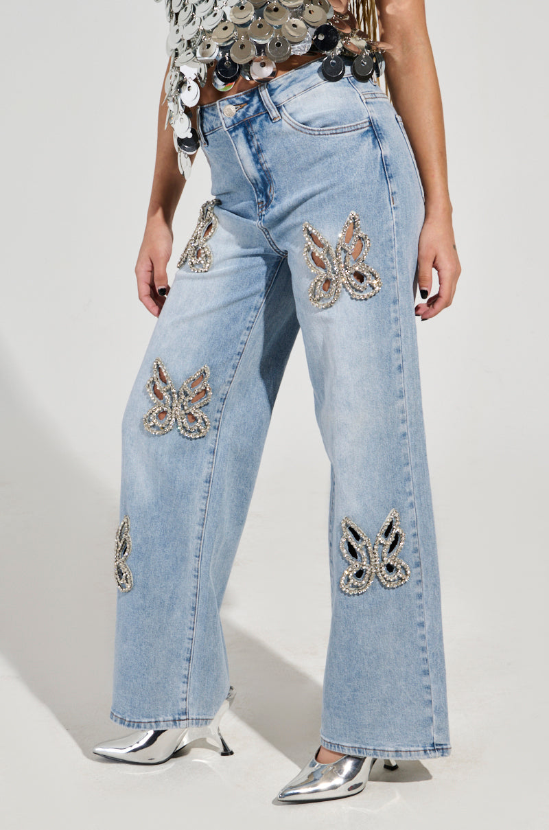 A person is dressed in light blue RHINESTONE BUTTERFLY CUTOUT DENIM JEANS that offer a Y2K-inspired wide-leg style. They enhance their outfit with metallic silver shoes and a top embellished with large silver sequins, set against a plain white background.
