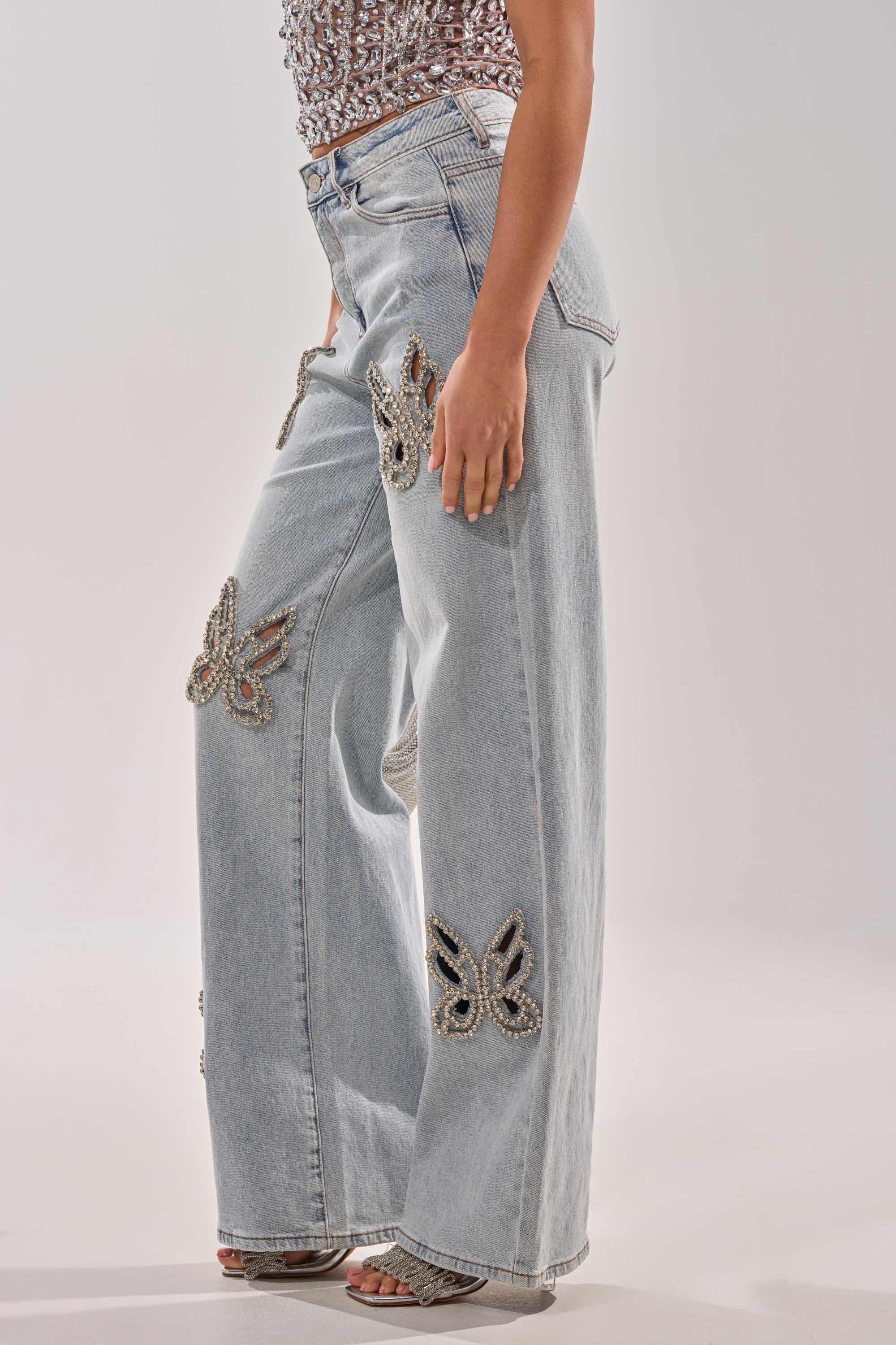 A person wears RHINESTONE BUTTERFLY CUTOUT DENIM JEANS in light blue, featuring large beaded butterfly appliqués, paired with a sequined sleeveless top and open-toe heels against a plain light background. 2