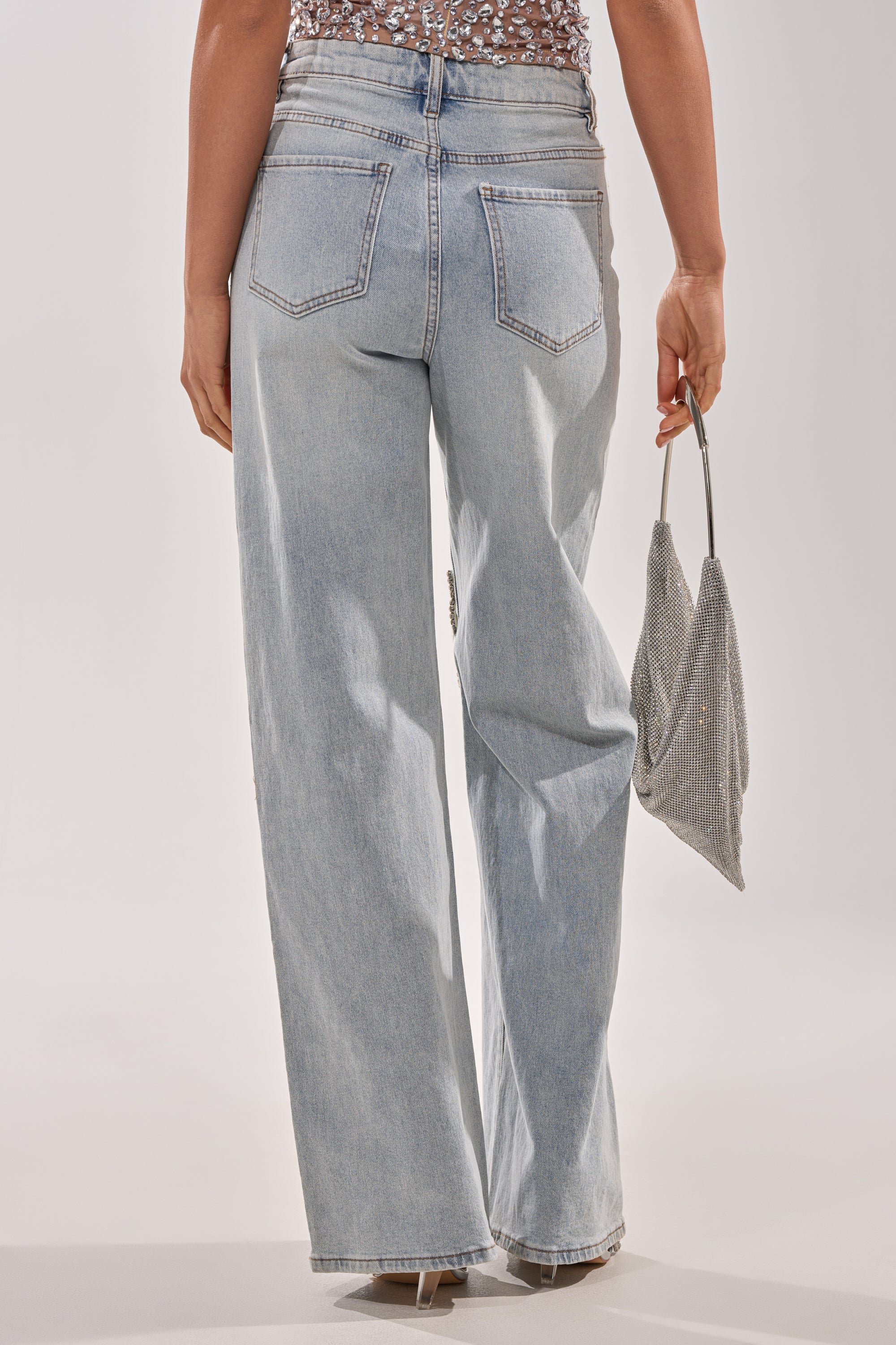 A person is shown from the back against a plain background, wearing RHINESTONE BUTTERFLY CUTOUT DENIM JEANS and holding a small, sparkly silver handbag, channeling effortless Y2K style. 3
