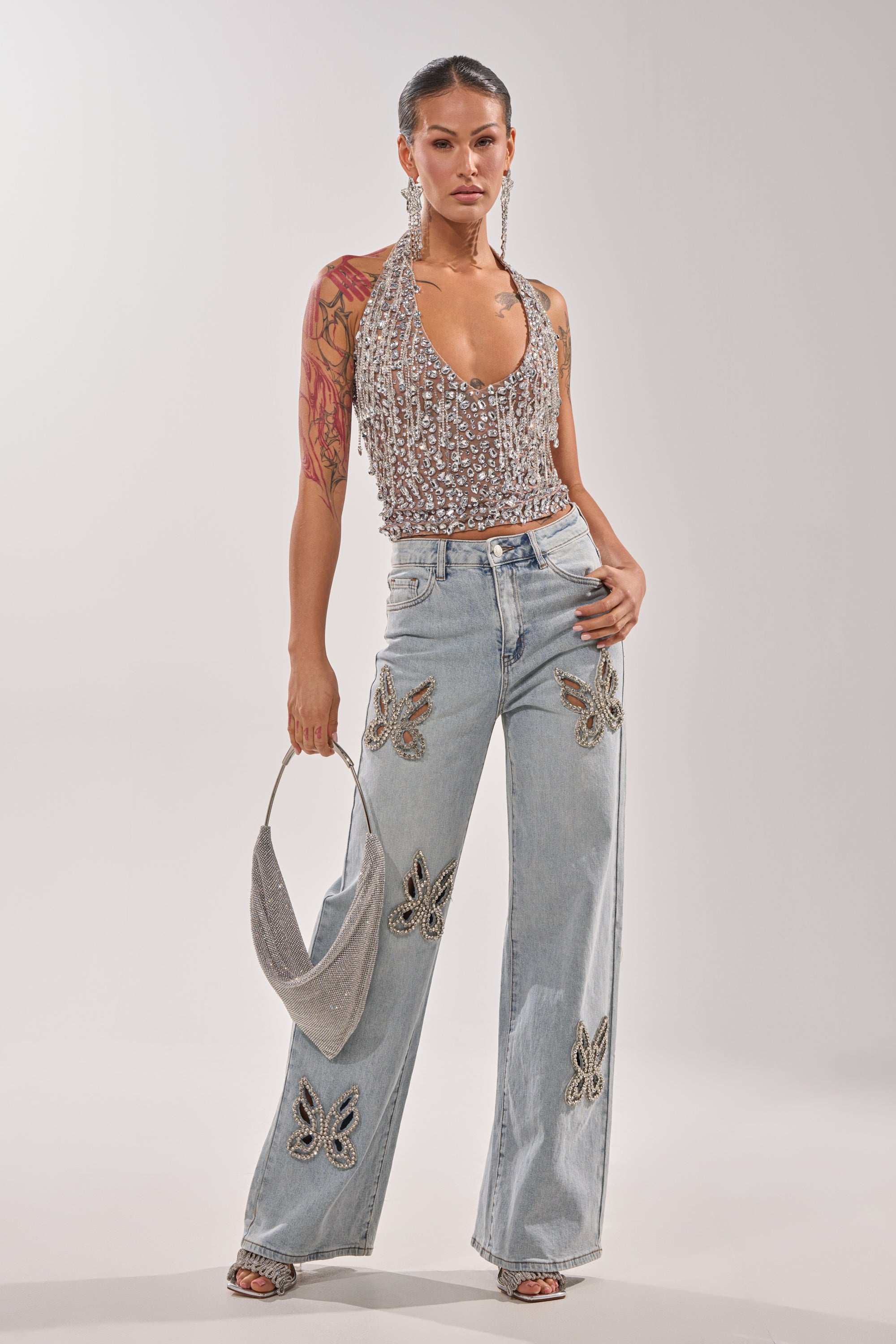 A woman exudes Y2K vibes in RHINESTONE BUTTERFLY CUTOUT DENIM JEANS with butterfly details, a sparkly halter top, dangling earrings, silver handbag, tattooed arm, and slicked-back hair. 5