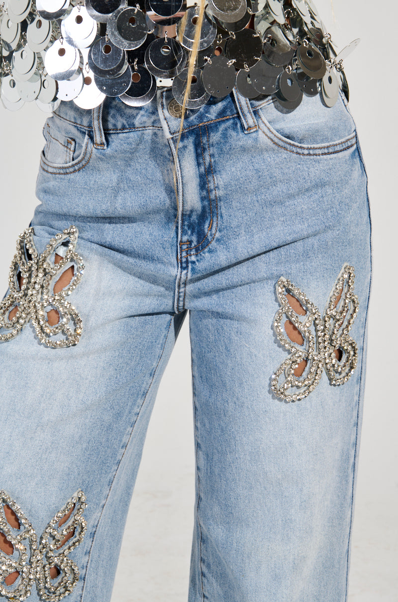 A close-up of a person wearing RHINESTONE BUTTERFLY CUTOUT DENIM JEANS, inspired by Y2K fashion. The top part of the individual showcases a silver sequin top, shimmering above the waistline of these statement jeans.
