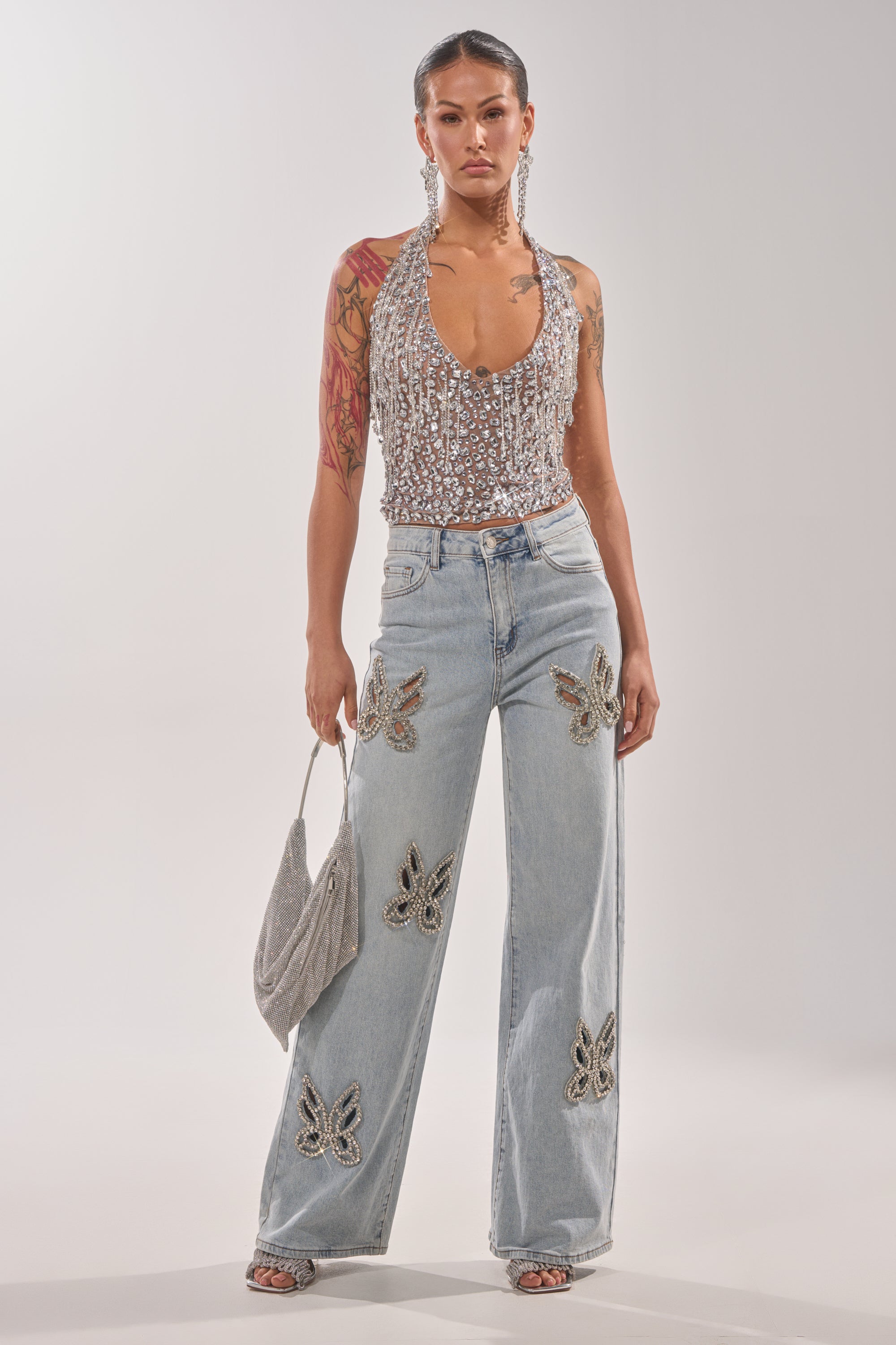 A woman stands confidently in a sparkly halter top, RHINESTONE BUTTERFLY CUTOUT DENIM JEANS, statement earrings, and a glittery handbag, with visible tattoos on her left arm. 6