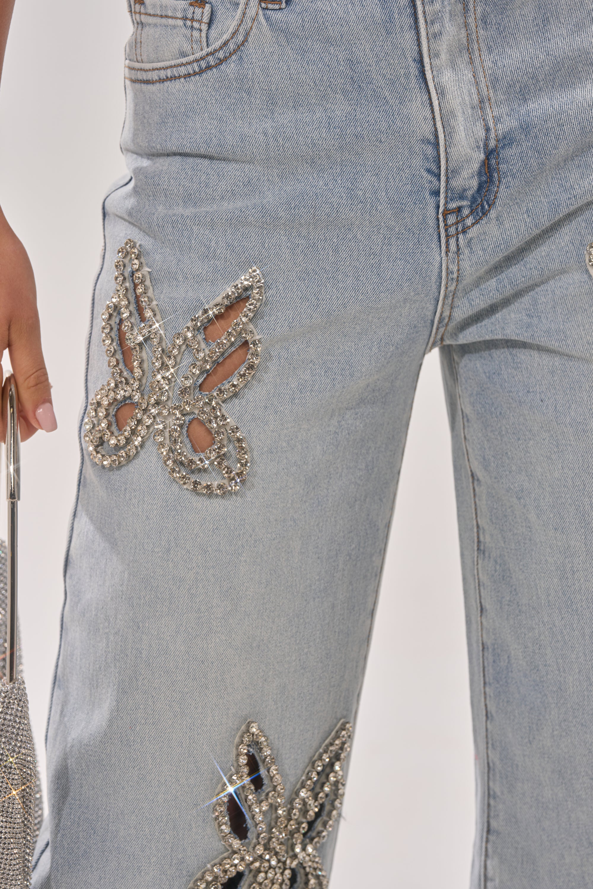Close-up of RHINESTONE BUTTERFLY CUTOUT DENIM JEANS with sparkling rhinestone butterfly designs. A person holds a matching shiny bag on the left, highlighting the trendy Y2K-inspired denim style. 7