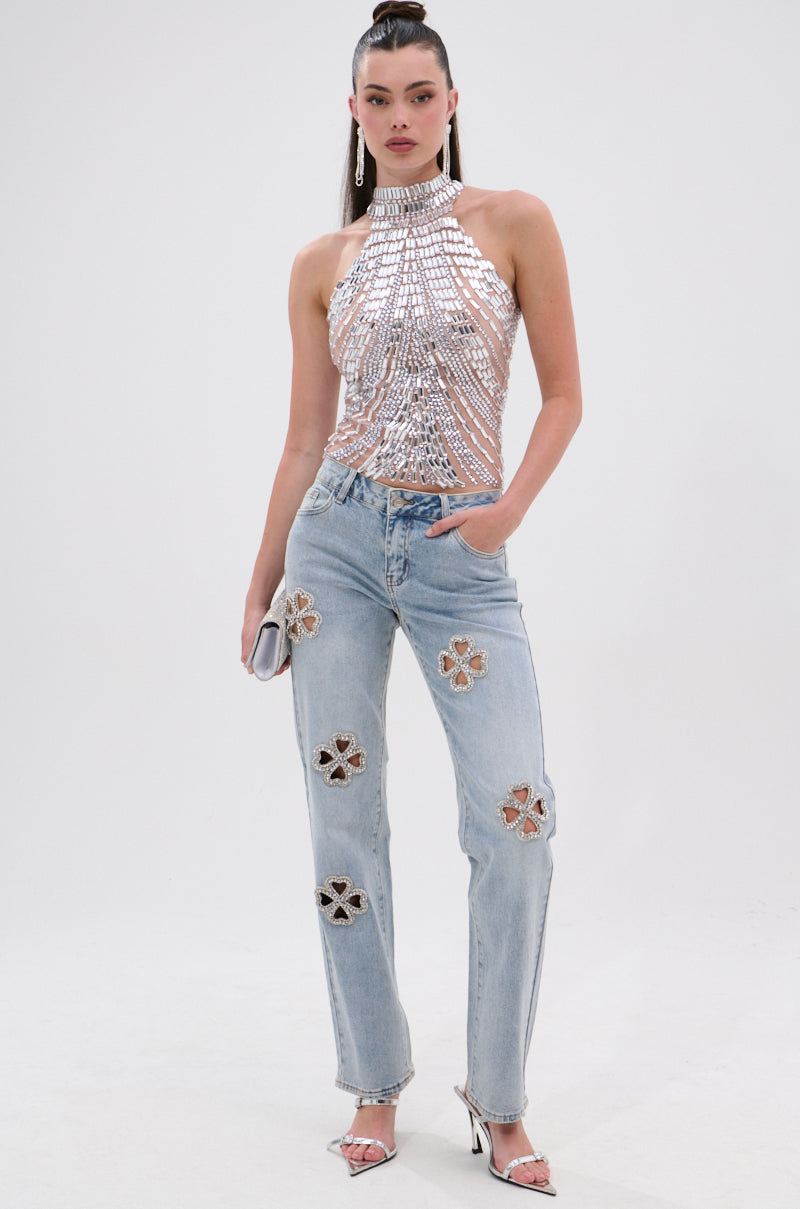 A woman poses against a white background wearing sparkling silver heels, drop earrings, a shiny halter top, a small silver clutch, and DIVINE FEMININE ENERGY RHINESTONE DETAIL JEANS with cut-out floral patches.