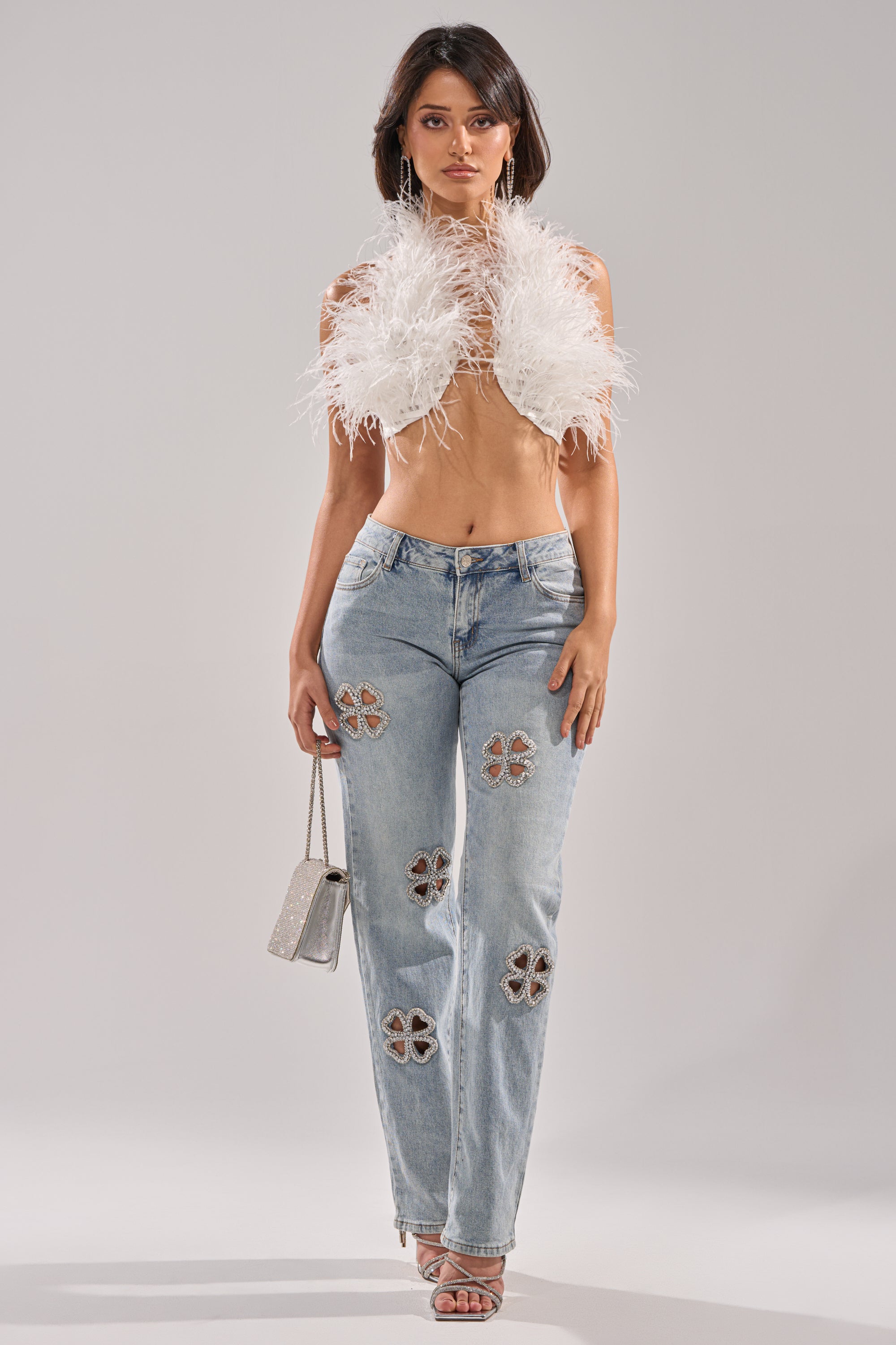 A woman poses against a plain background wearing a white feathered crop top, silver heels, a small silver handbag, and DIVINE FEMININE ENERGY RHINESTONE DETAIL JEANS in a light wash with clover cut-out details.