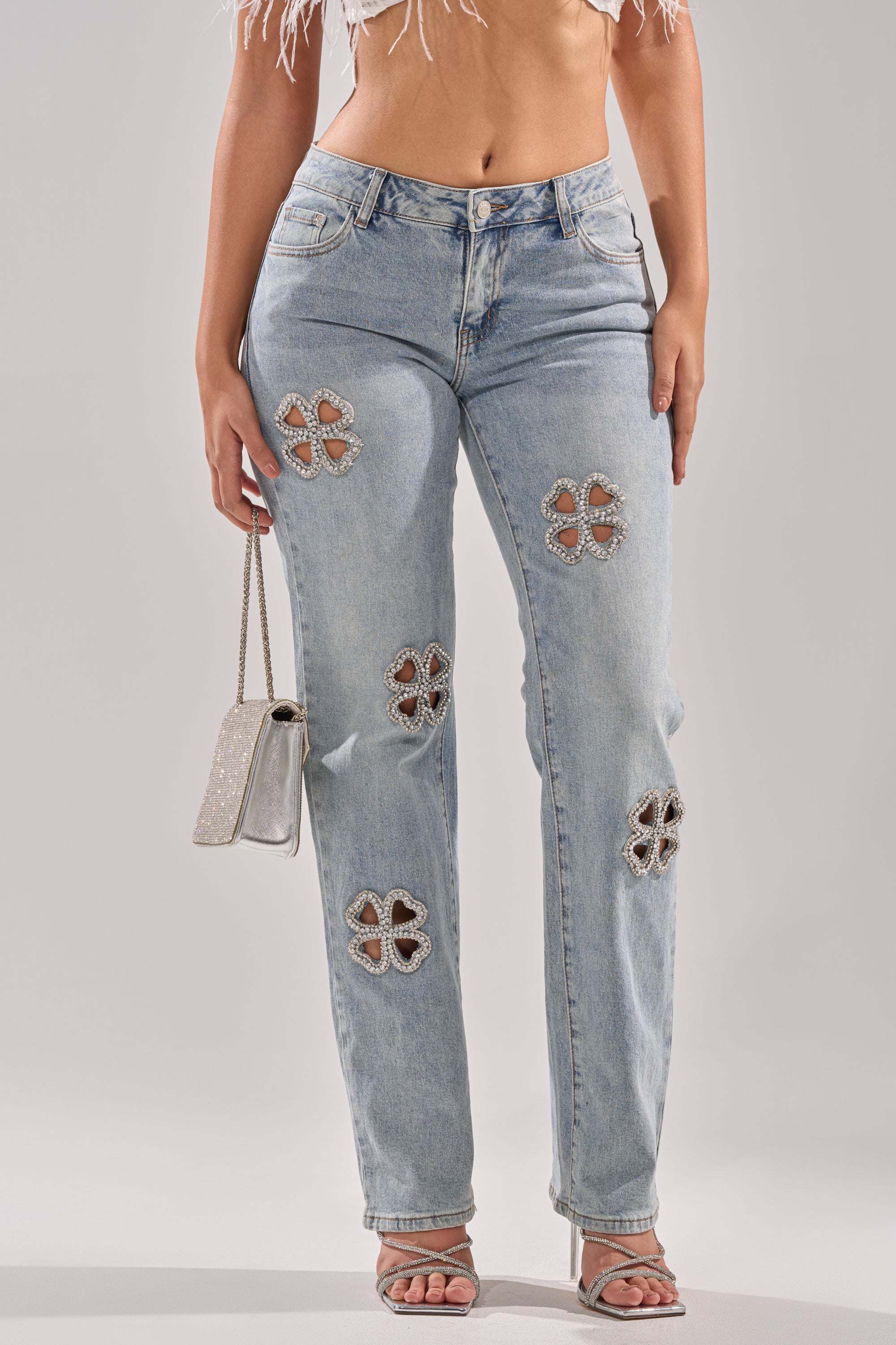 A woman wears DIVINE FEMININE ENERGY RHINESTONE DETAIL JEANS, a white crop top, high-heeled sandals, and carries a small silver purse.