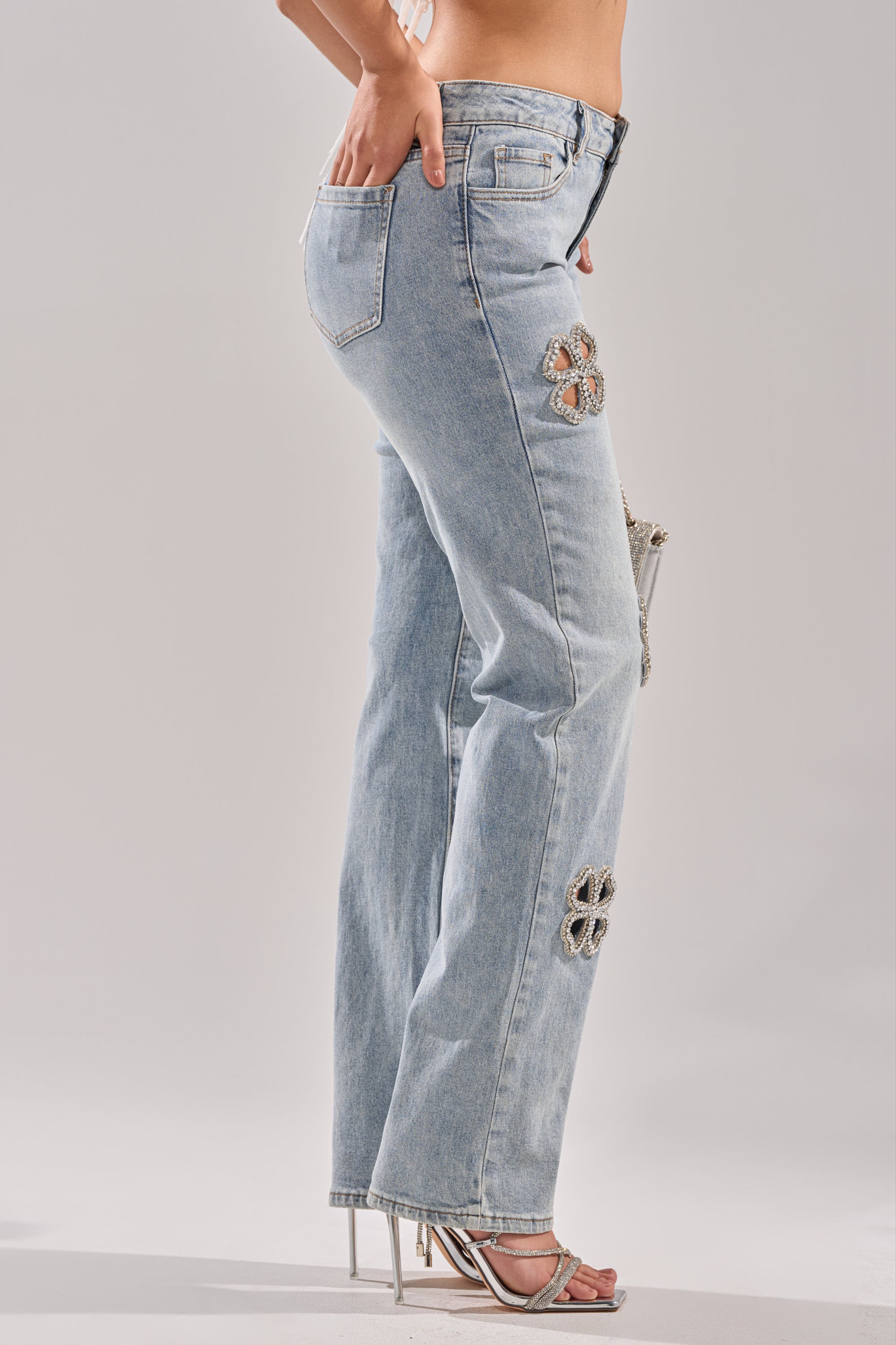 Wearing DIVINE FEMININE ENERGY RHINESTONE DETAIL JEANS—light blue, high-waisted with flower rhinestone embellishments on the side—and silver strappy high-heeled sandals, shown from the side against a plain background.