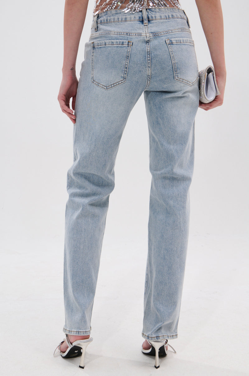 A person faces away against a white background, wearing DIVINE FEMININE ENERGY RHINESTONE DETAIL JEANS in a light wash with straight legs, white heeled sandals, and holding a small clutch in their right hand.