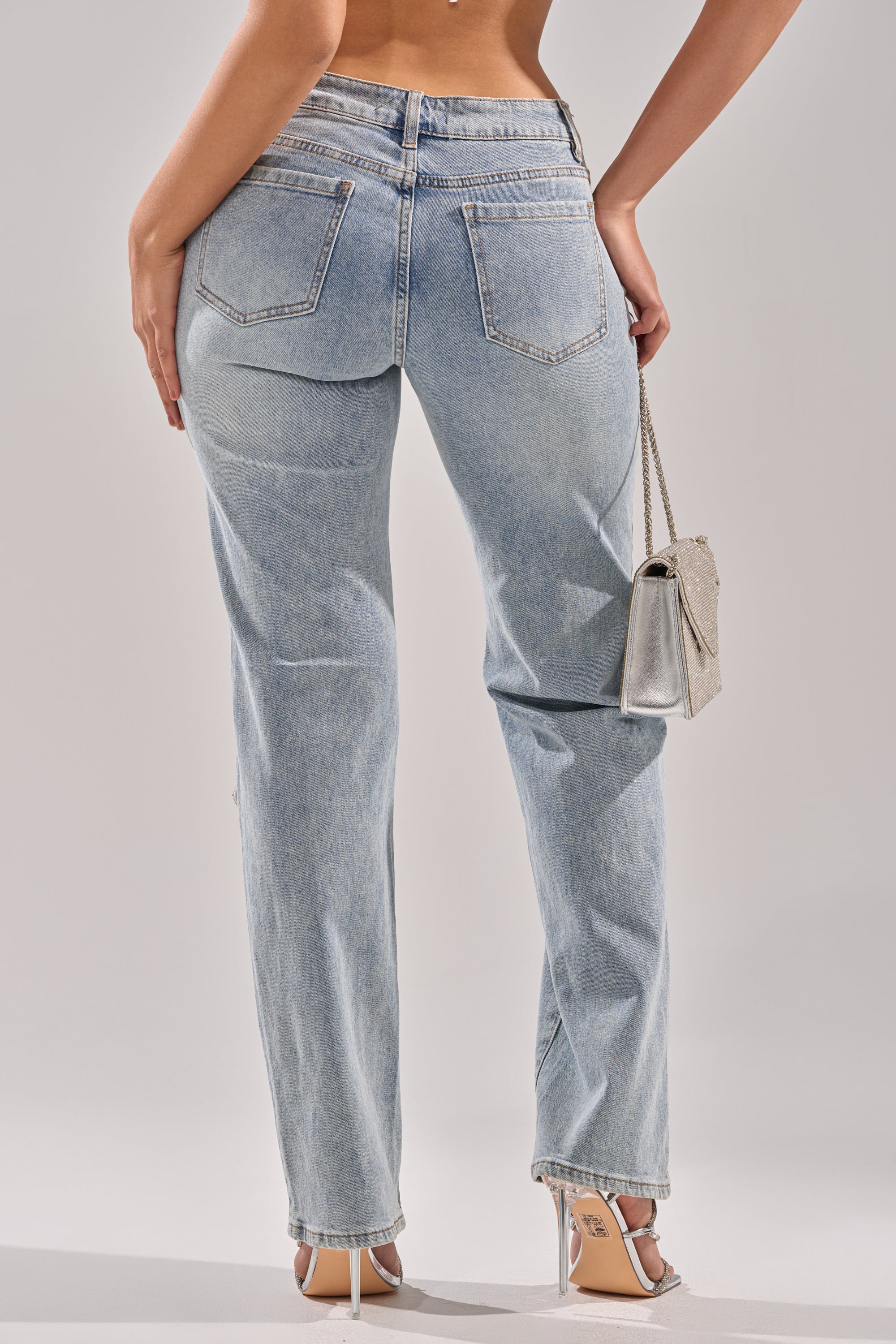 A person wearing DIVINE FEMININE ENERGY RHINESTONE DETAIL JEANS, styled with beige high-heeled sandals and a small beige handbag. Shot from behind, the image highlights the jeans' back pockets and straight-leg fit.