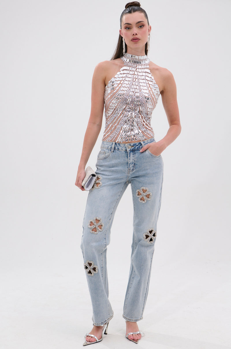 Radiating divine feminine energy, a woman wears silver sequin halter, DIVINE FEMININE ENERGY RHINESTONE DETAIL JEANS with star cutouts, silver heels, and a small clutch against a plain white background.