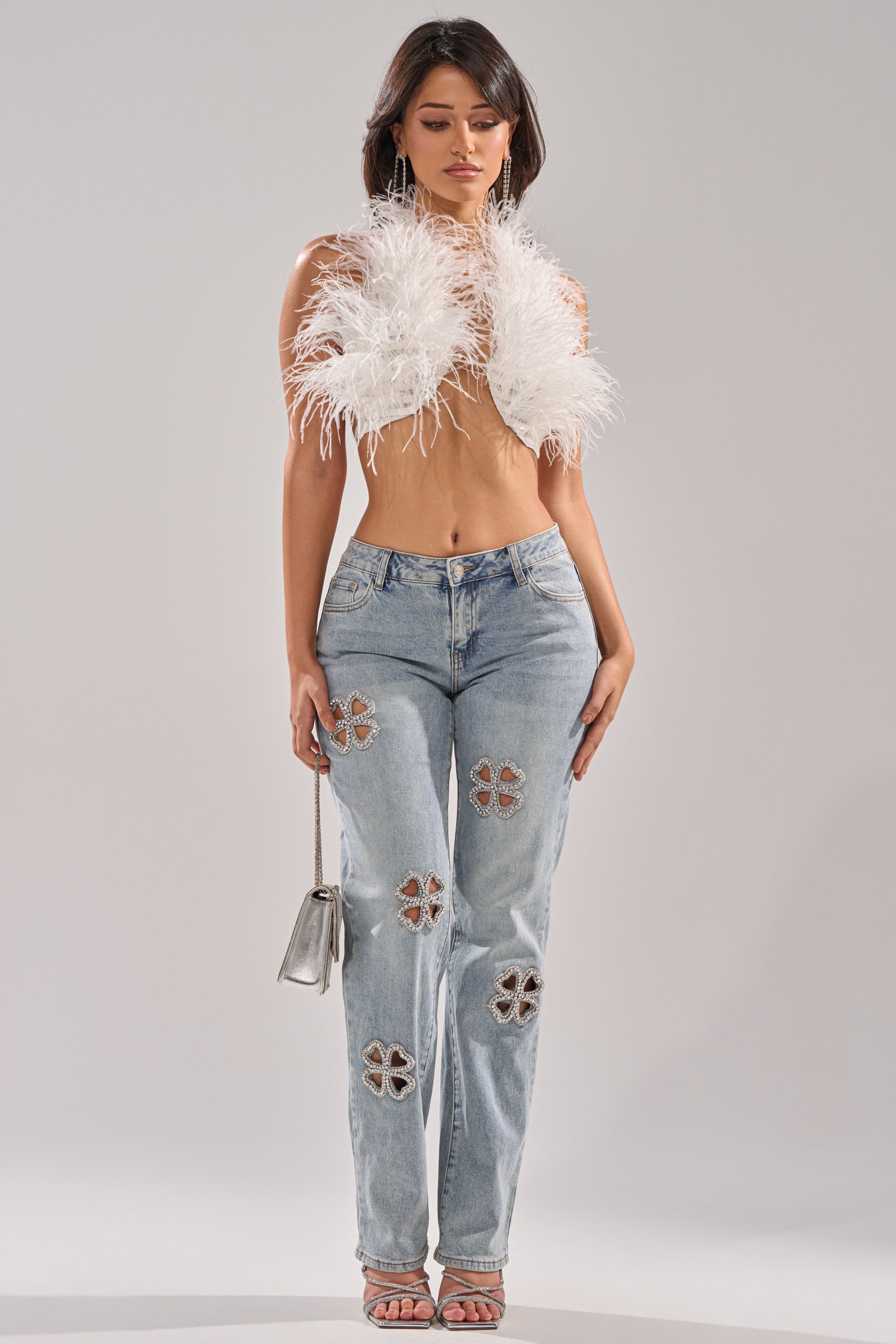 A woman faces forward in DIVINE FEMININE ENERGY RHINESTONE DETAIL JEANS, a white feathered crop top, silver sandals, and holding a small silver purse against a plain gray background.